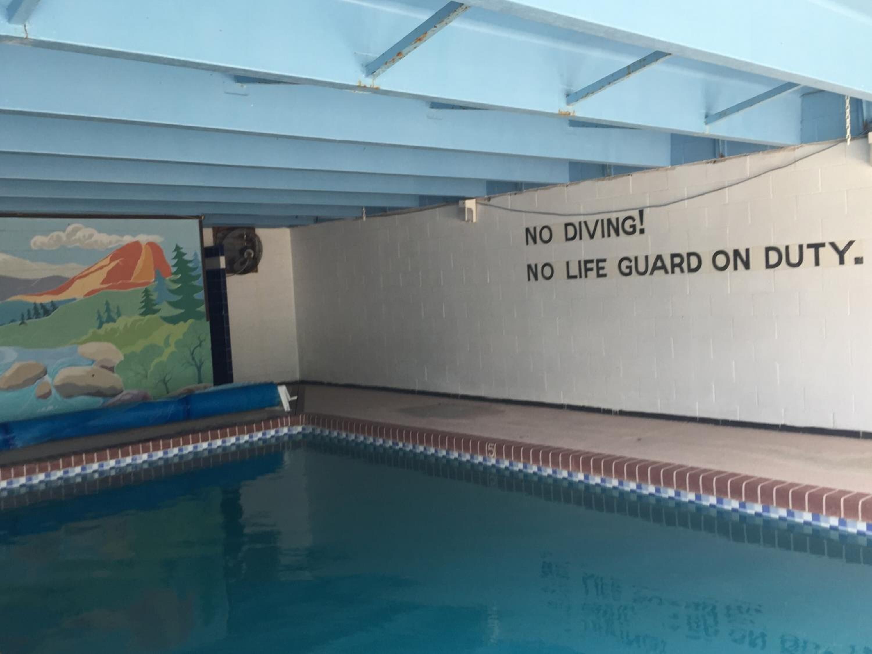 An indoor swimming pool with a mural of a mountain landscape and a warning sign stating "NO DIVING! NO LIFE GUARD ON DUTY."