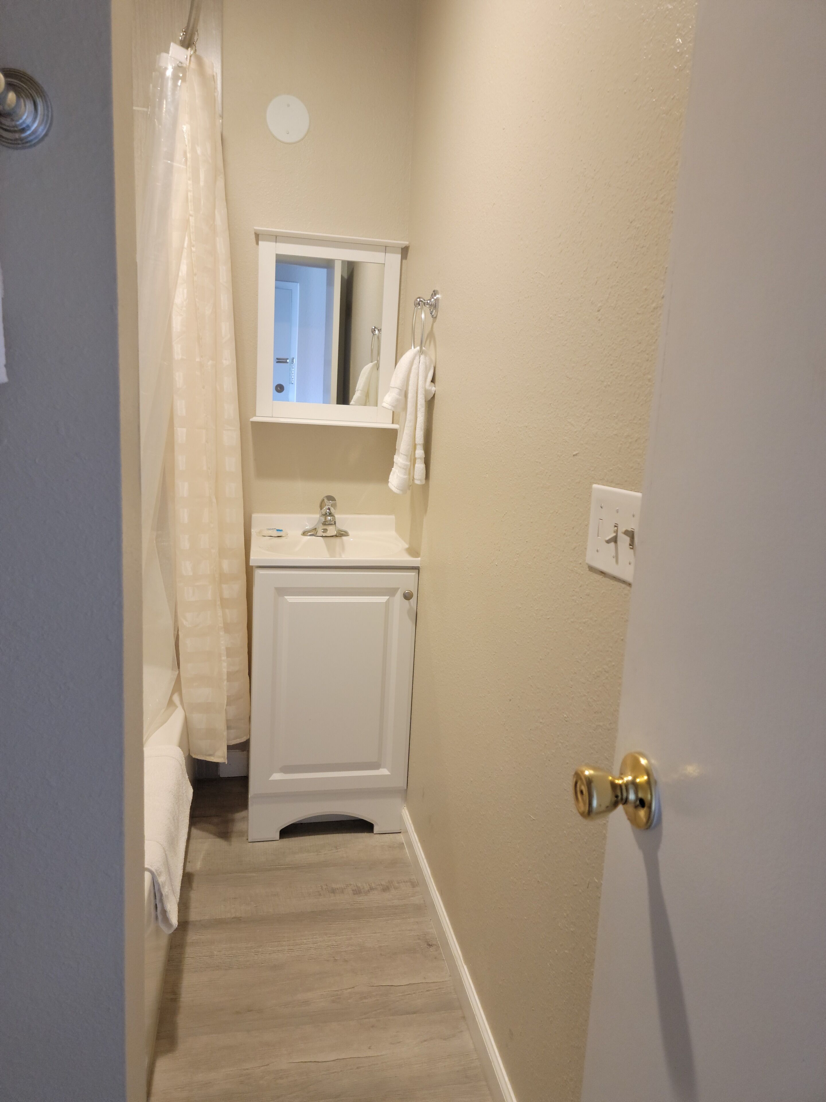 A narrow bathroom with a white toilet, a sink, and a mirror above it. There is a towel hanging on the wall and a shower curtain partially visible.