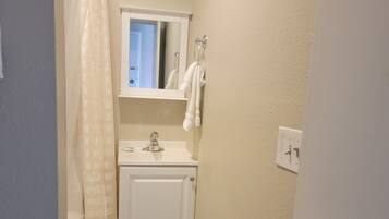 A narrow bathroom with a white toilet, a sink, and a mirror above it. There is a towel hanging on the wall and a shower curtain partially visible.