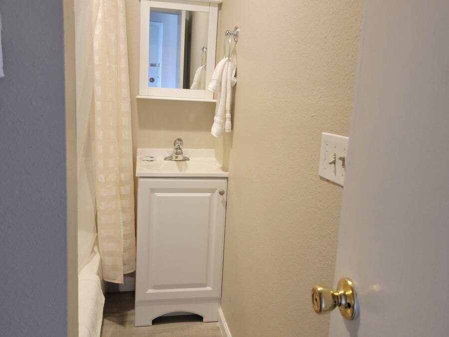 A narrow bathroom with a white toilet, a sink, and a mirror above it. There is a towel hanging on the wall and a shower curtain partially visible.