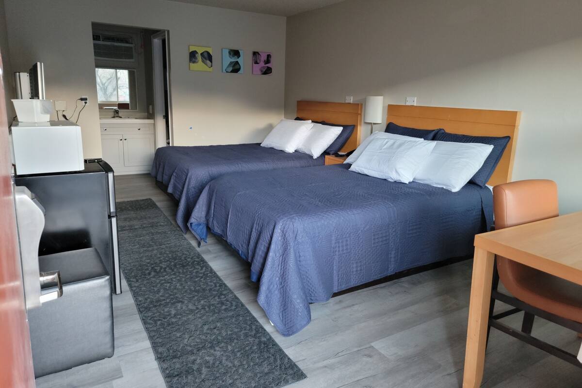 Une chambre d’hôtel avec deux lits, un bureau, une chaise, une petite table, une lampe de bureau, un climatiseur fixé au mur et une fenêtre.