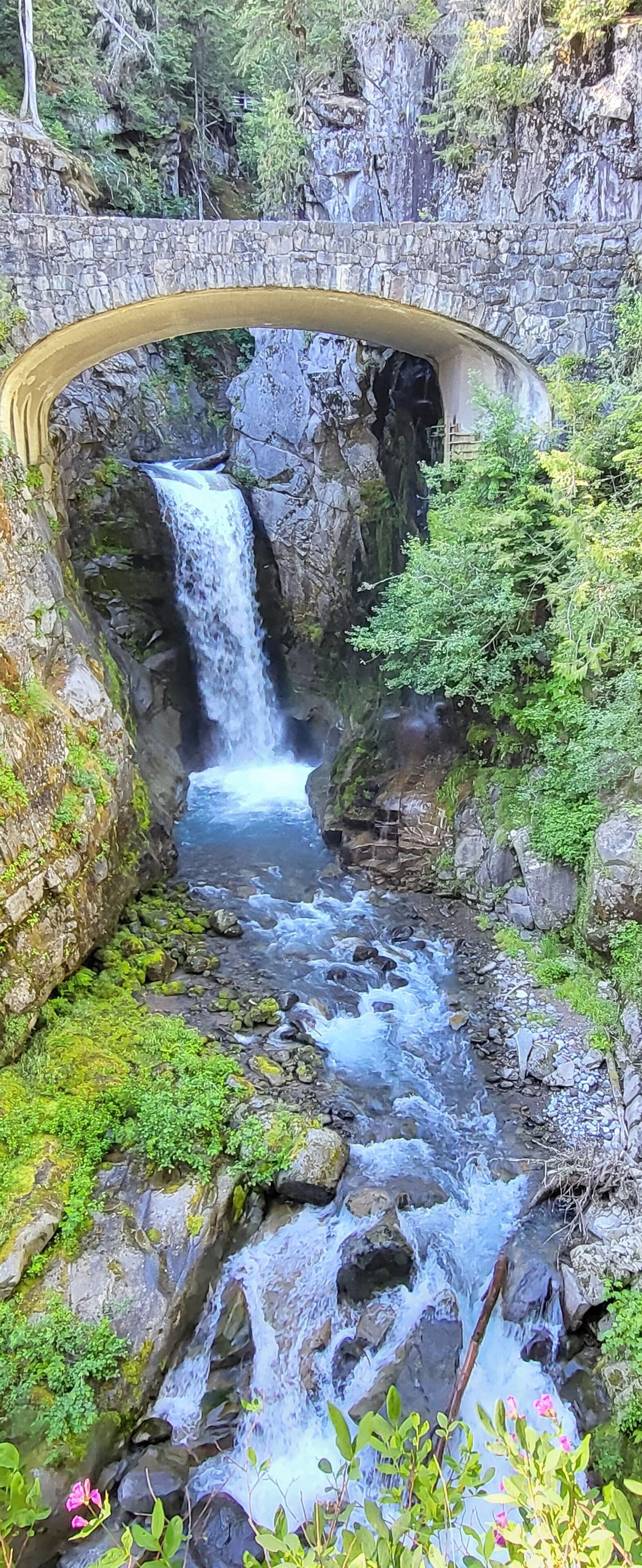 A stone bridge arching over a cascading waterfall surrounded by lush greenery.