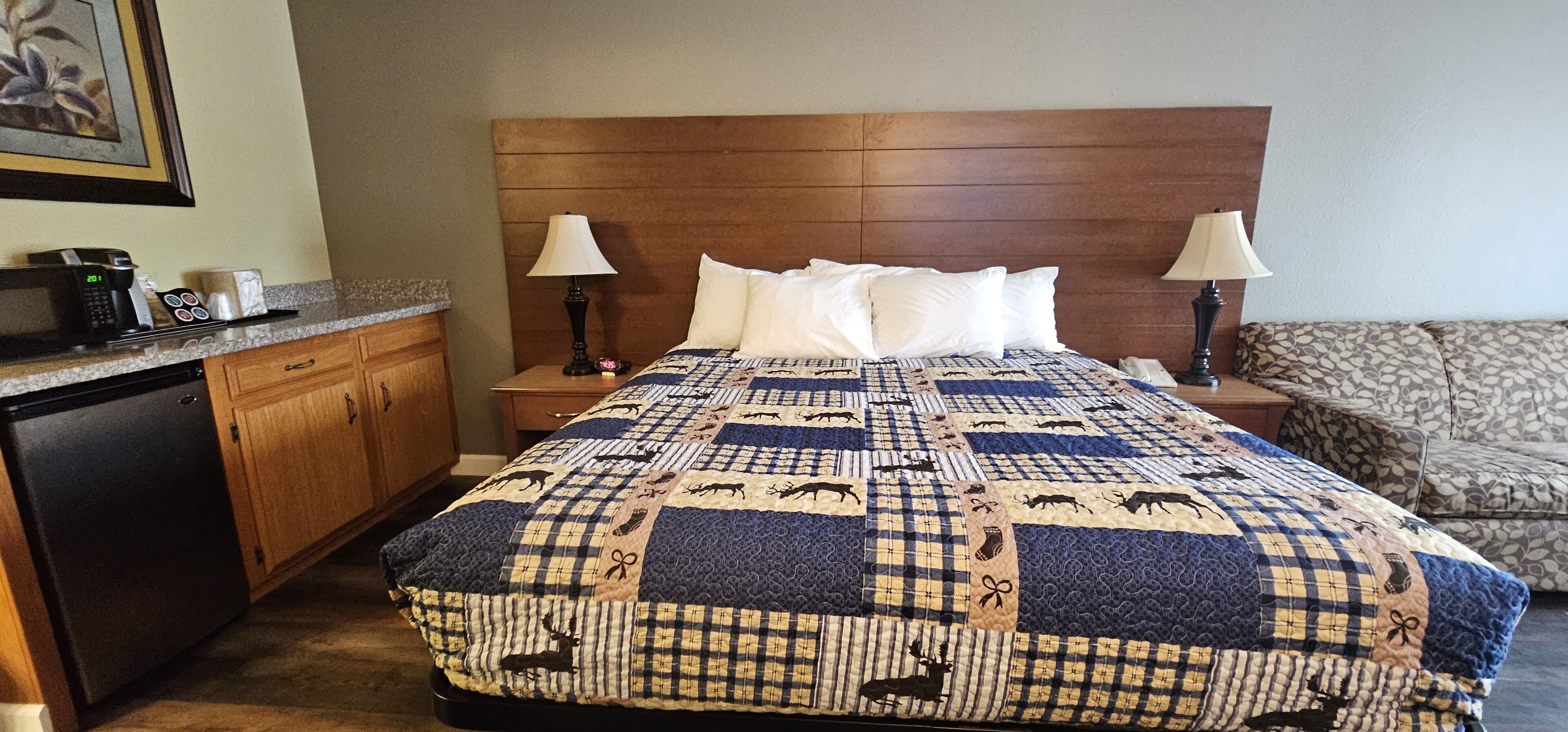 Habitación de hotel con una cama grande, cabecera de madera, dos lámparas de noche, una mesita de noche con un teléfono, un microondas y un refrigerador pequeño.