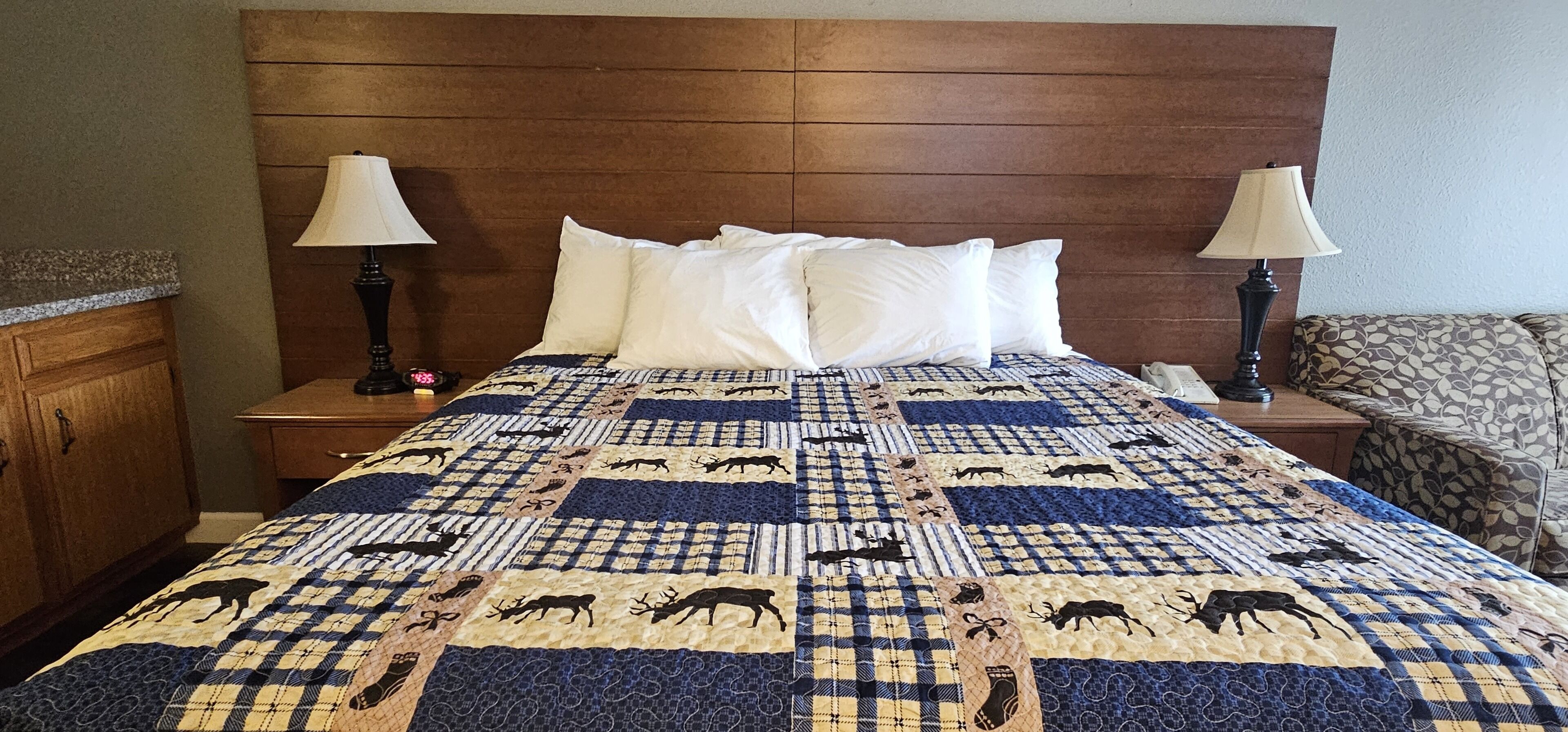 A hotel room with a large bed, wooden headboard, two bedside tables with lamps, a patterned quilt, and a patterned armchair.
