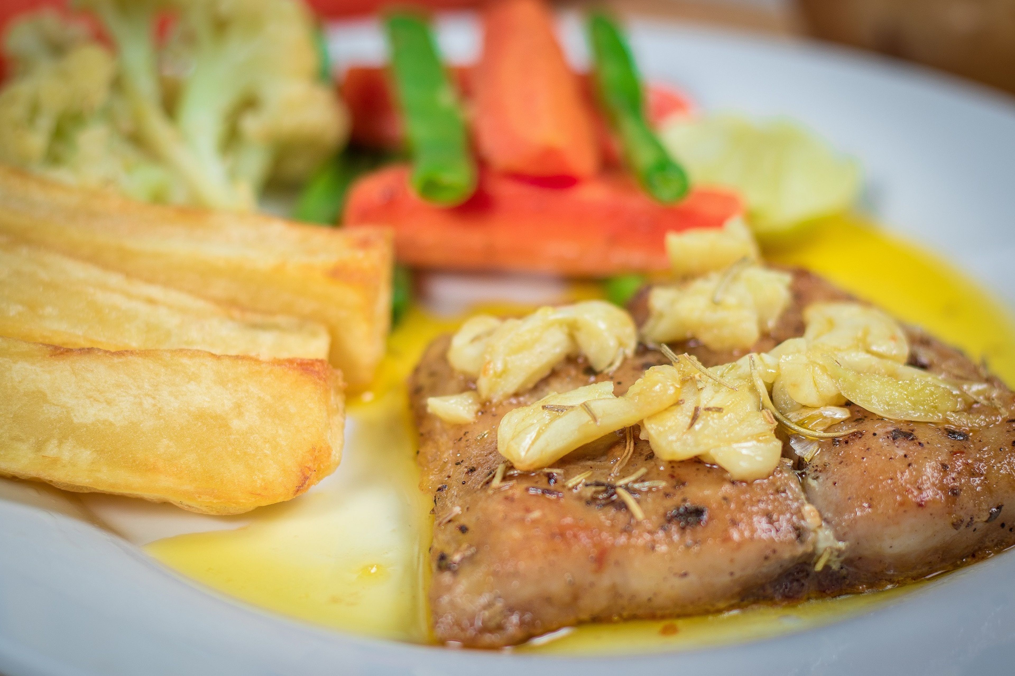 Une assiette contenant un morceau de poisson garni d’ail et d’herbes, accompagné de frites et d’une salade.
