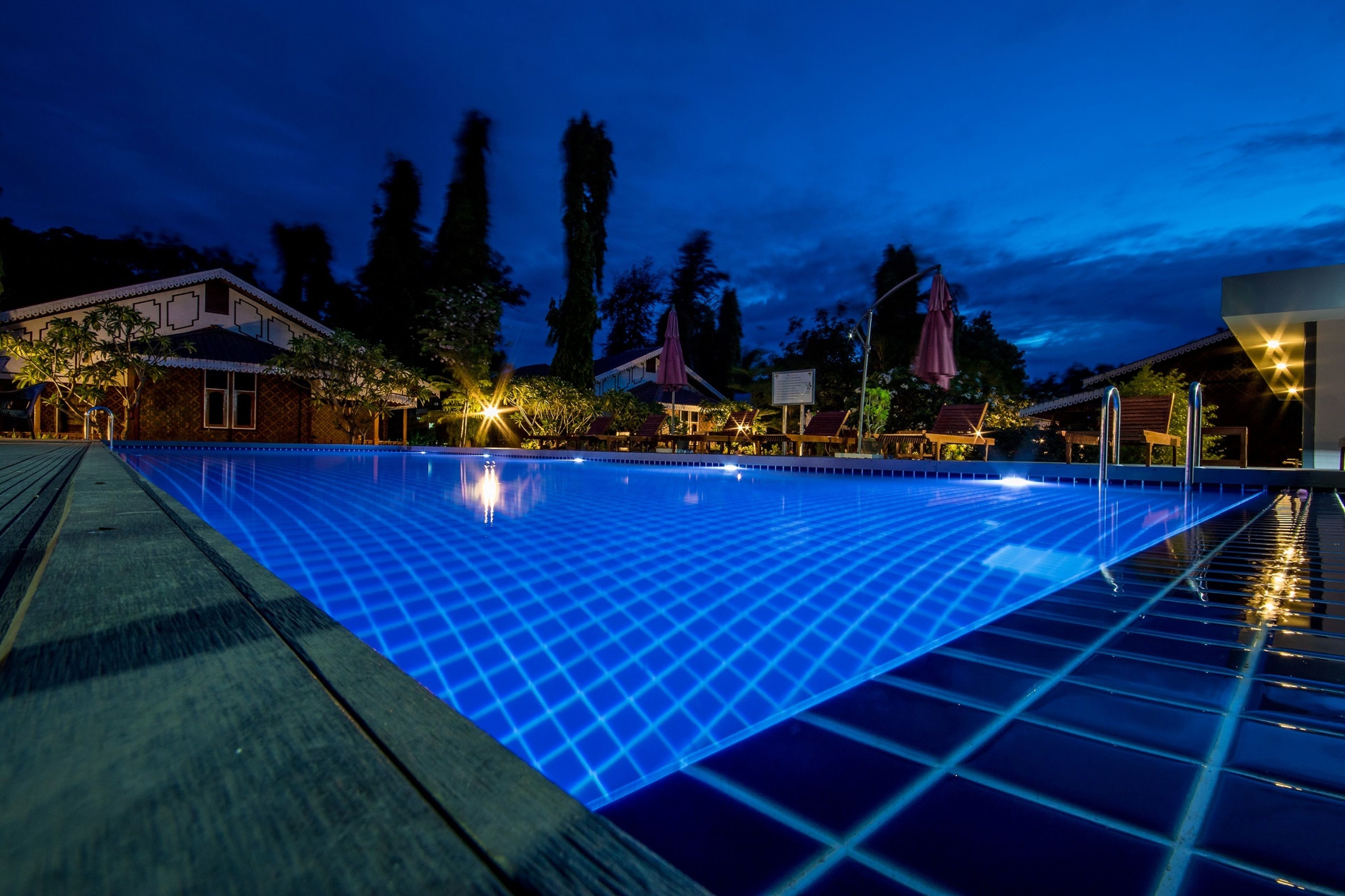 Una piscina al anochecer con un patrón de luces en forma de cuadrícula, rodeada de edificios y árboles.