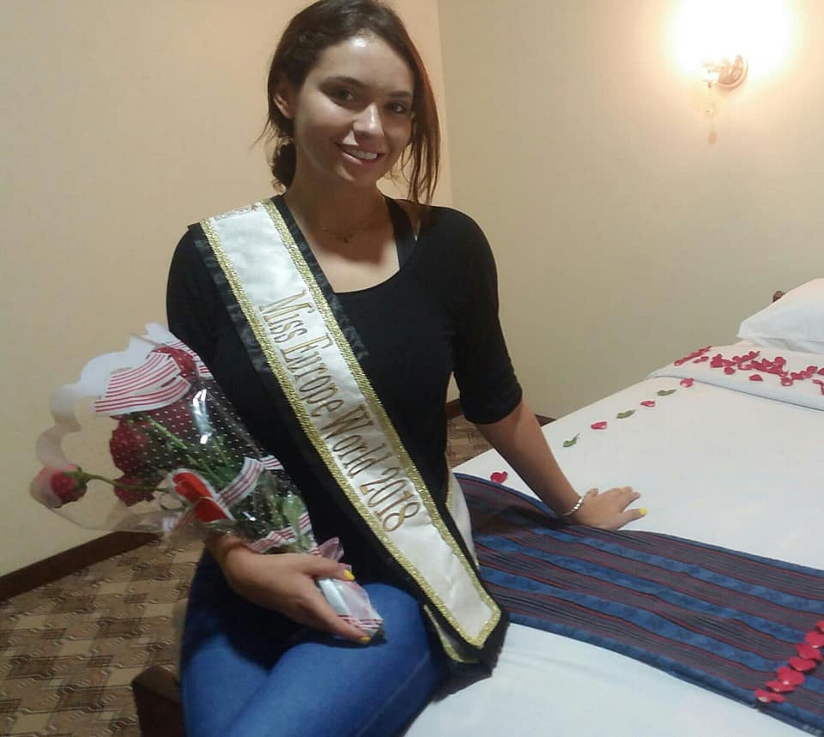 A person sitting on a bed with a sash that reads "Miss Europe World 2018".