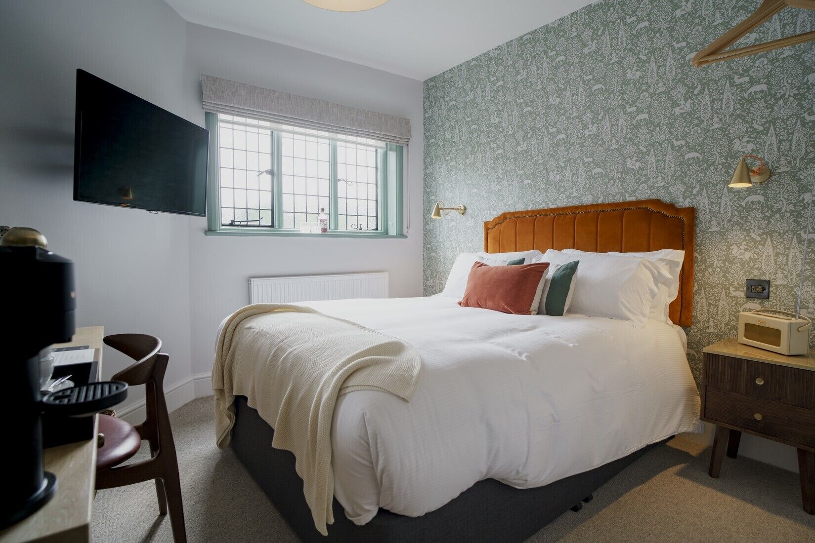 A bedroom with a bed, a desk, a chair, a radiator, a window, and a wall-mounted TV.