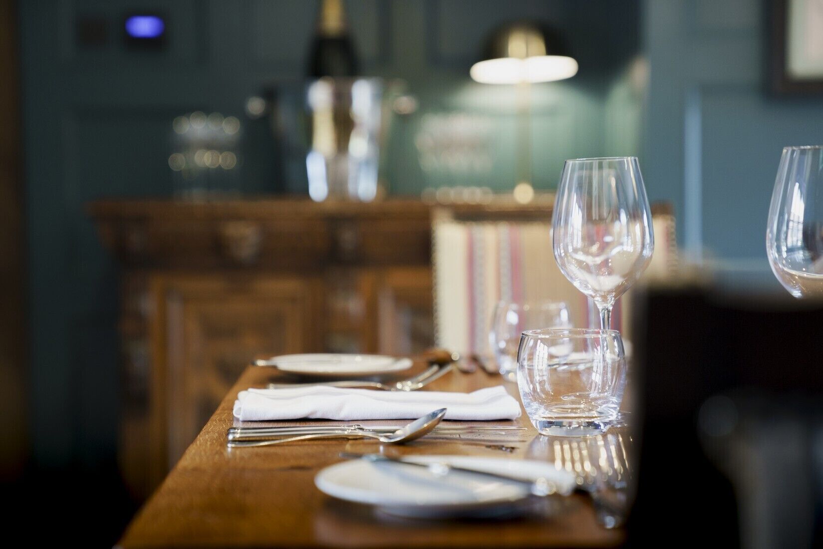 A dining table set with plates, cutlery, and glasses, with a wooden backdrop and a blurred background.