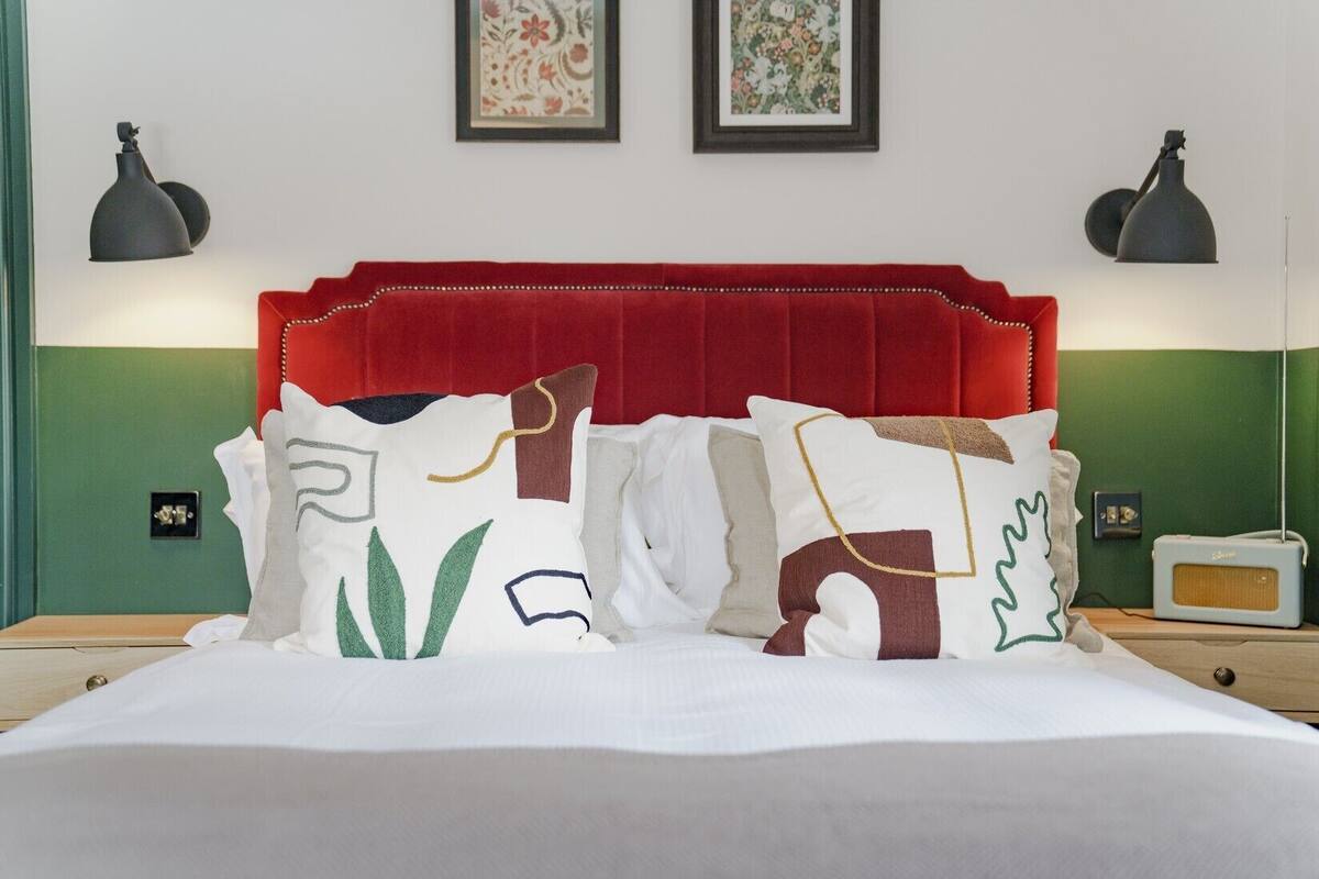 Una cama con cabecera roja, ropa de cama blanca y almohadas con estampados geométricos coloridos. Hay dos lámparas de pared y dos obras de arte con motivos florales enmarcadas sobre la cama.