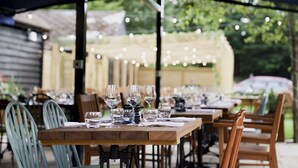 Un área de comedor al aire libre con mesas dispuestas para dos, que presenta cristalería transparente y sillas de madera.