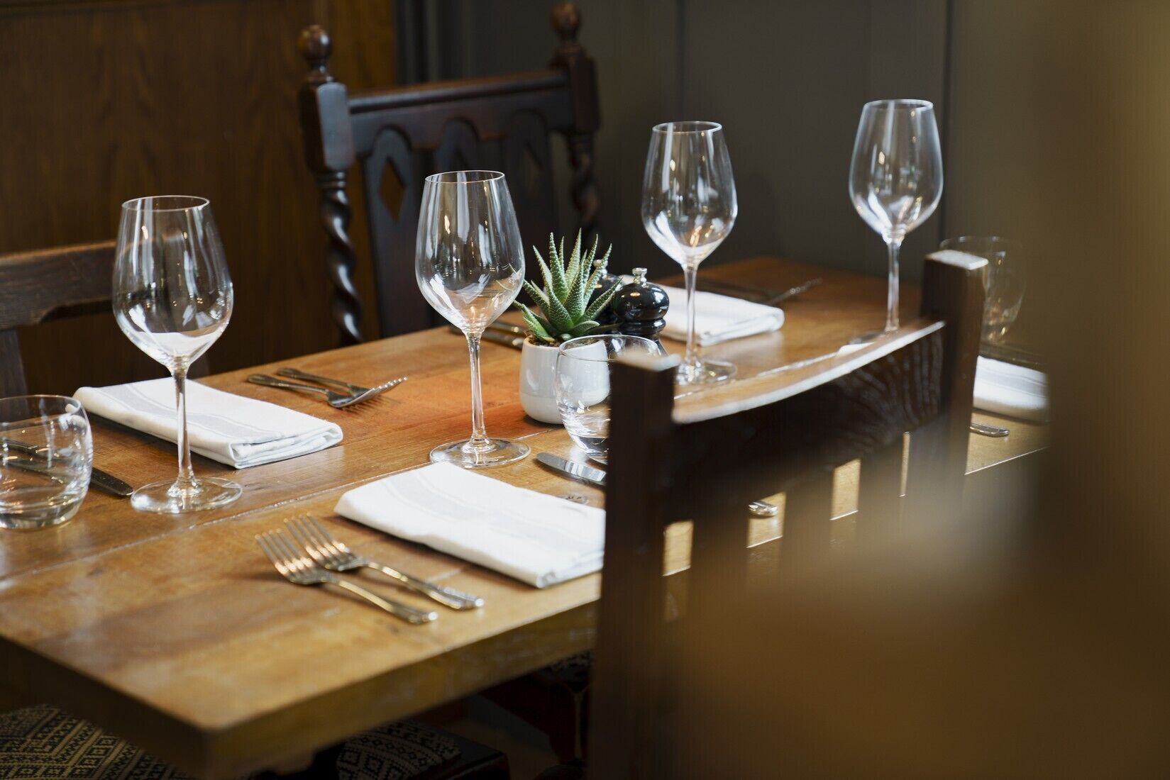 Una mesa de comedor lista con copas de vino, cubiertos y una pequeña planta en maceta.