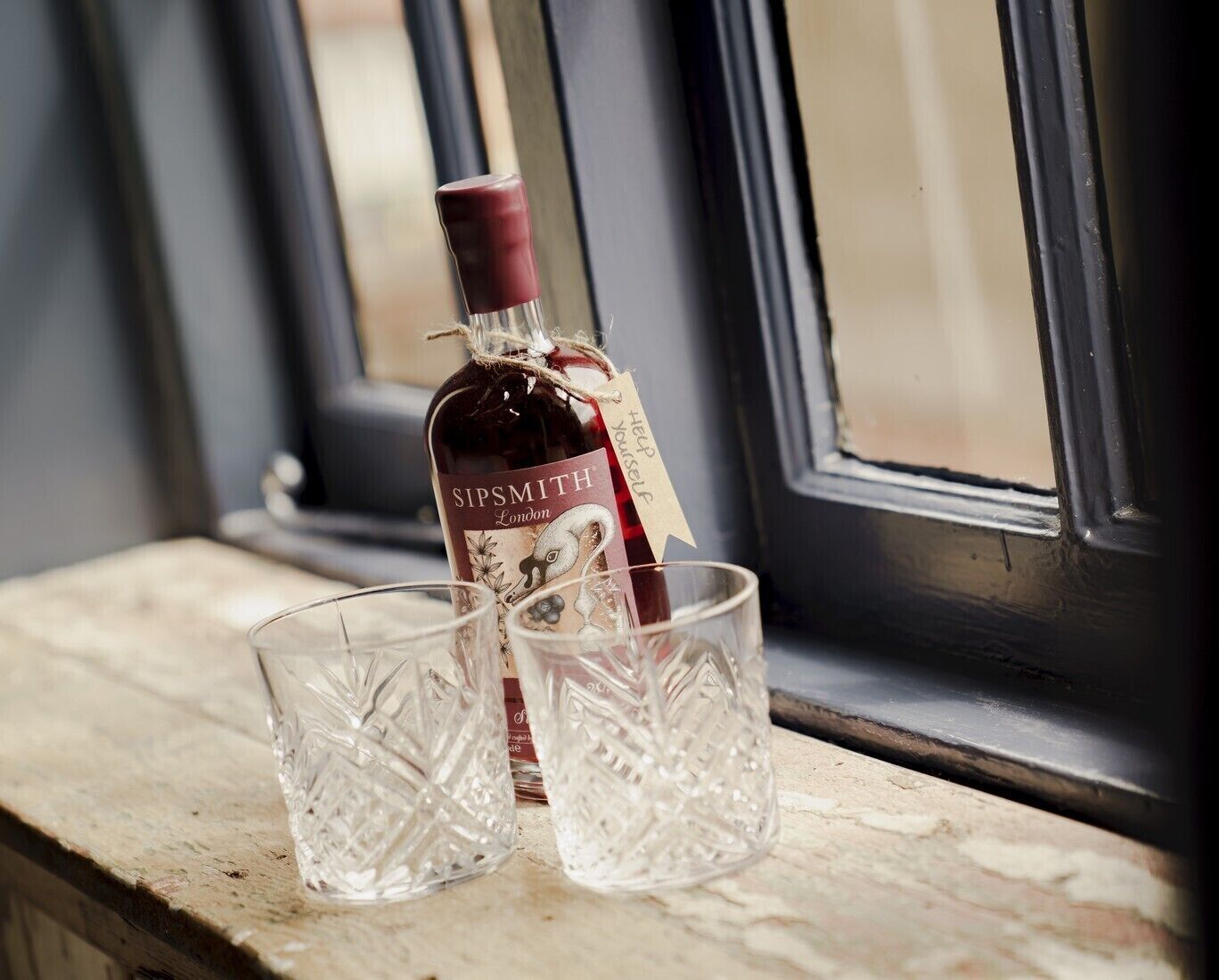 Uma garrafa de gin Sipsmith London e duas taças de cristal sobre um parapeito de madeira perto de uma janela.