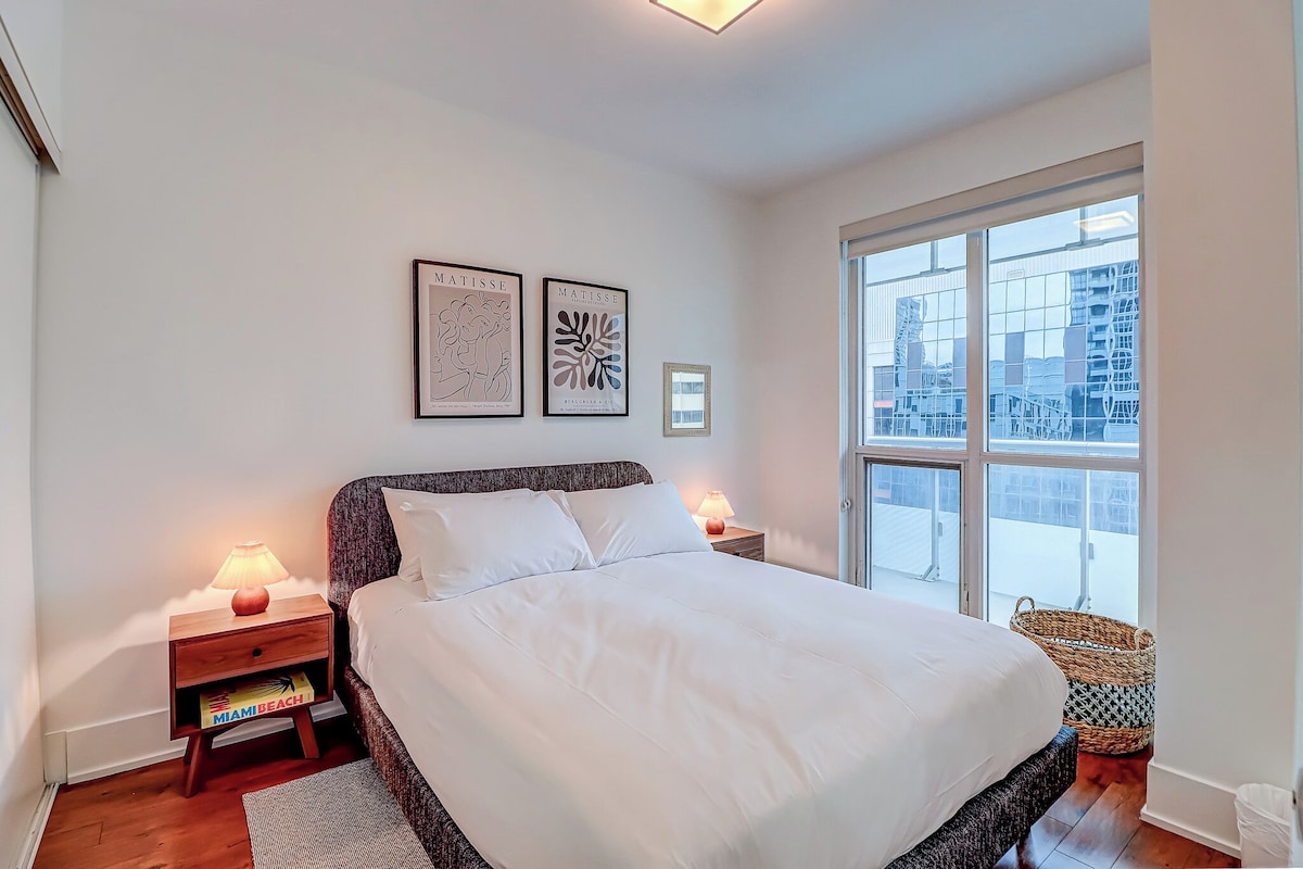Un dormitorio con una cama grande, dos mesitas de noche con lámparas, una ventana con vistas a edificios y dos cuadros enmarcados en la pared.