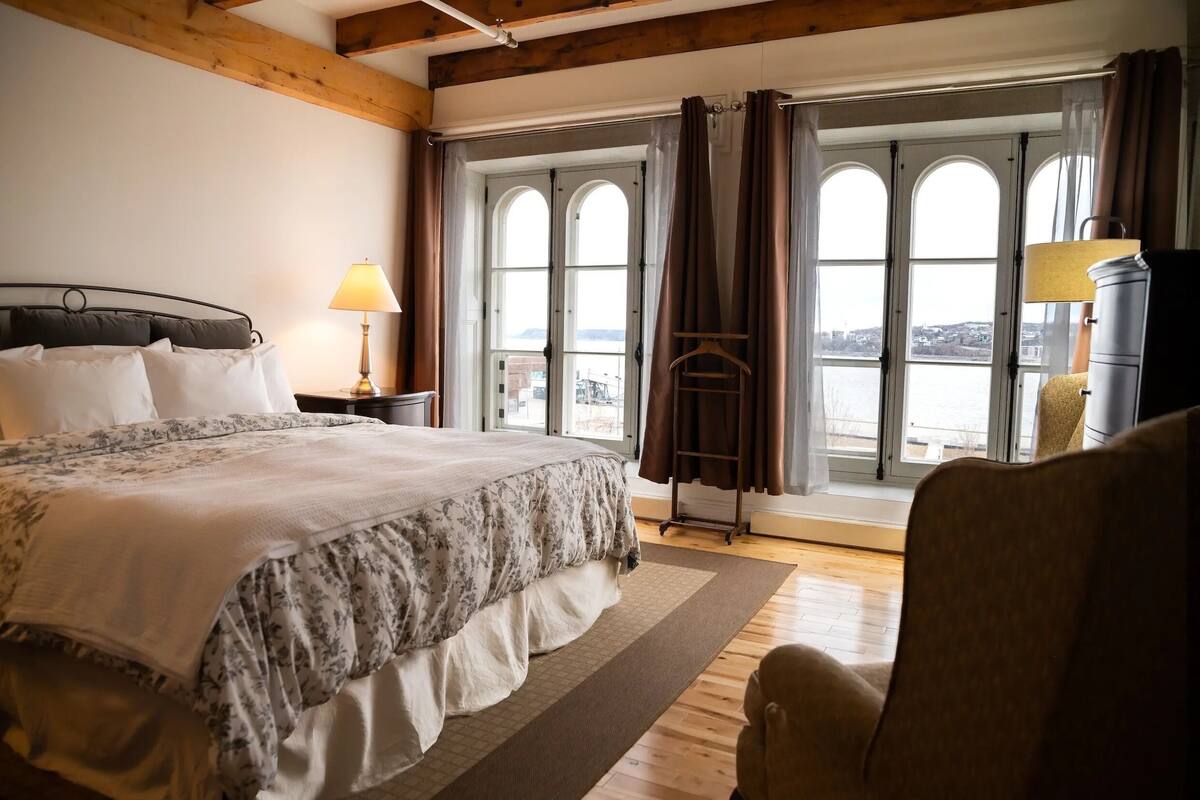 A bedroom with a large bed, a nightstand with a lamp, a chair, and a view of the water through large windows.