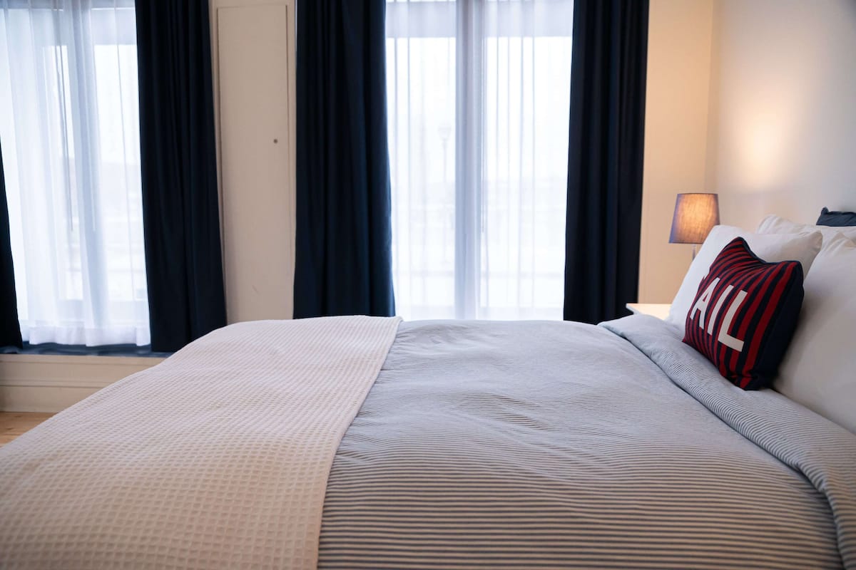 A neatly made bed with a striped pillow and a white blanket, a bedside lamp, and a window with sheer curtains.