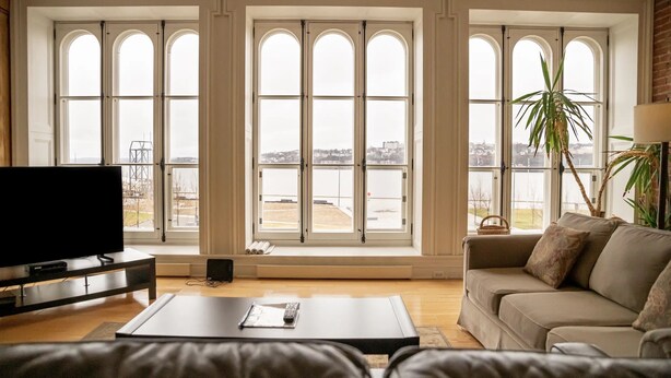 A spacious living room with large arched windows, a flat-screen TV, a sofa, and a coffee table. - Royal Dalhousie Condos (Québec City)