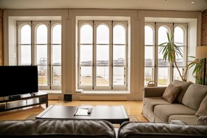 Een ruime woonkamer met grote boogvensters, een flatscreen televisie, een bank en een salontafel.