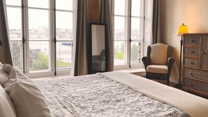 A hotel room with a large bed, a chair, a dresser, and a view of a cityscape through large arched windows. - Royal Dalhousie Condos (Québec City)