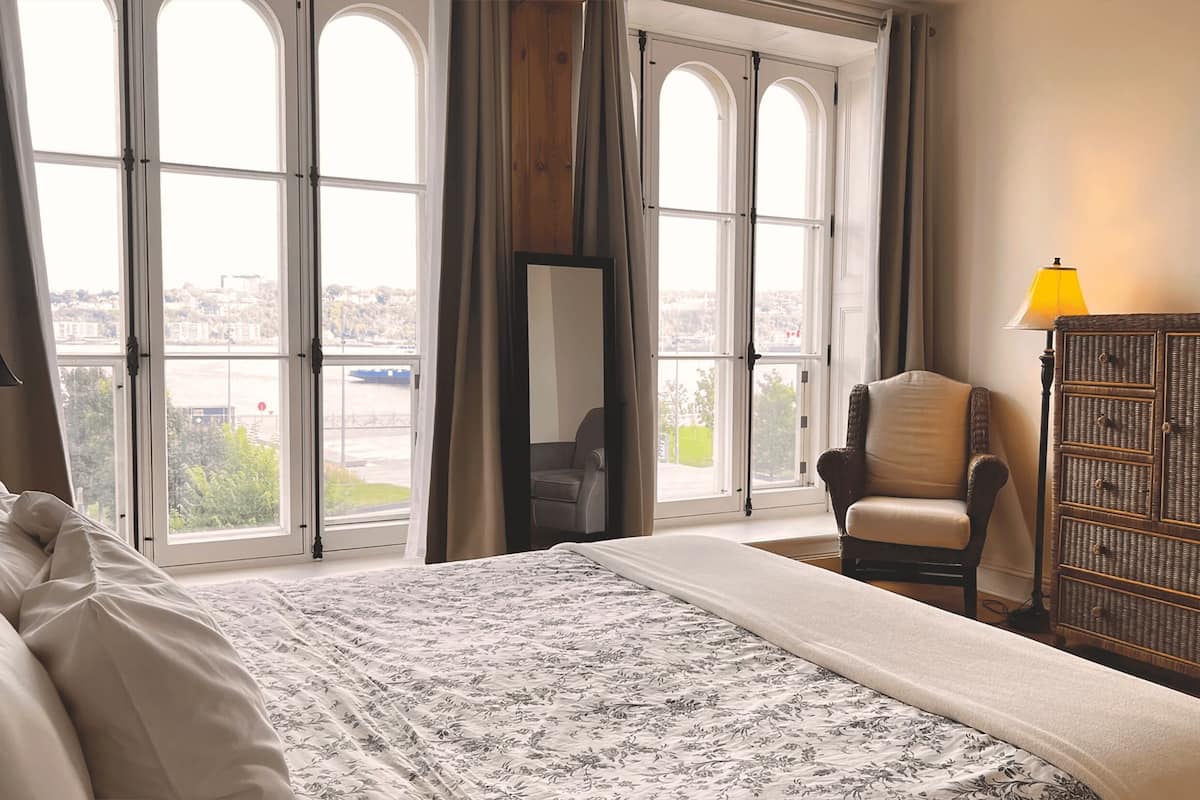 A hotel room with a large bed, a chair, a dresser, and a view of a cityscape through large arched windows.