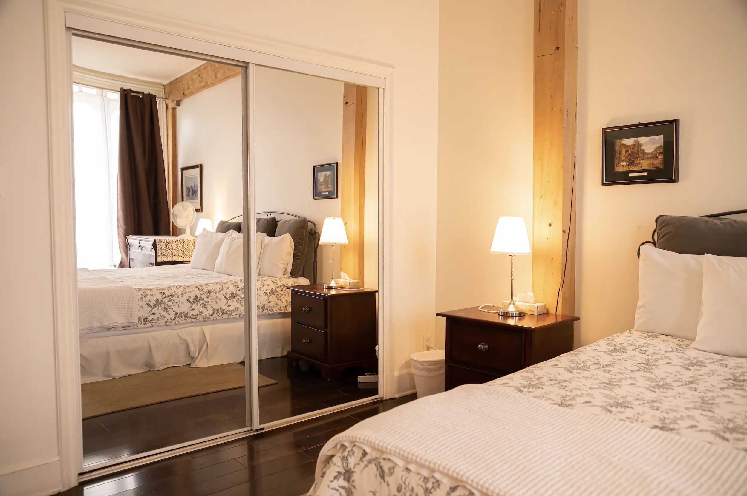A hotel room with a mirrored closet, a bed with a floral bedspread, a bedside table with a lamp, and a small bedside table with a lamp.