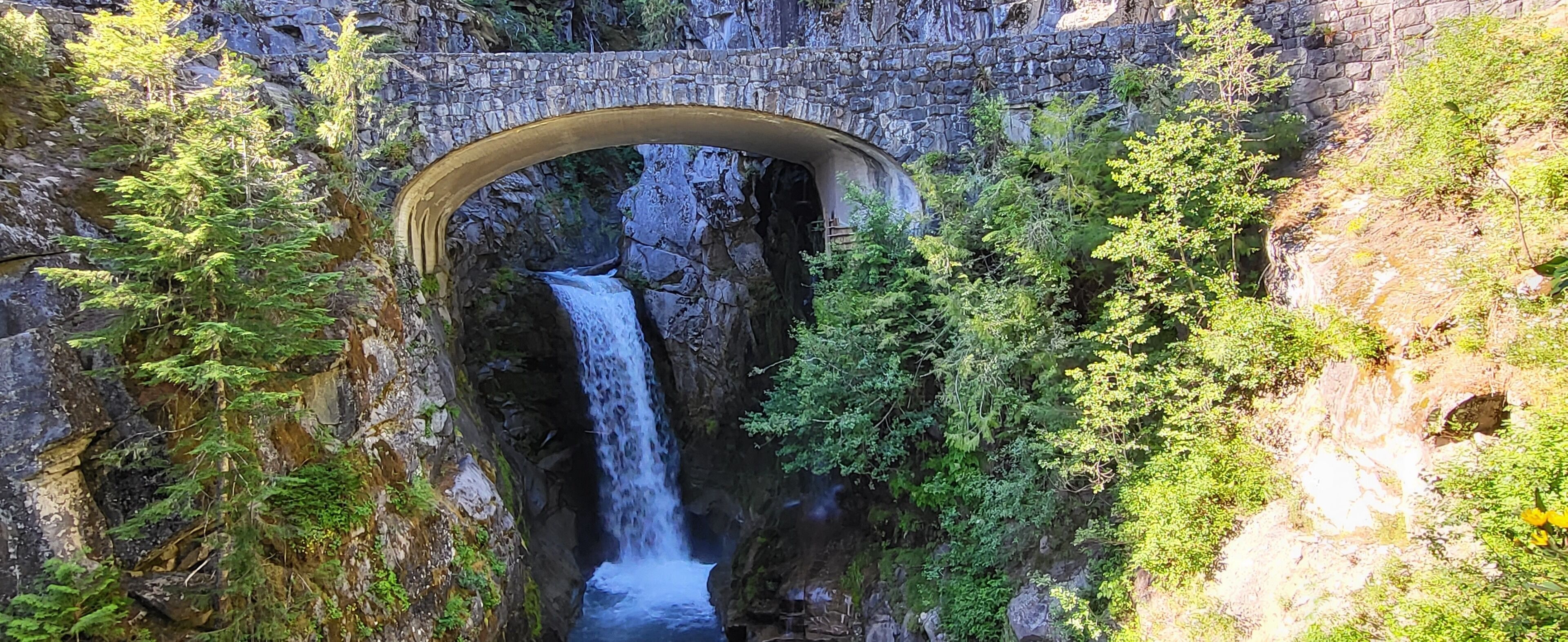 A stone bridge arching over a waterfall with lush greenery on either side.