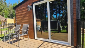 A wooden deck with two wicker chairs, a sliding glass door, and a view of a backyard with a lawn and trees. - Nisqually Lodge (Ashford)