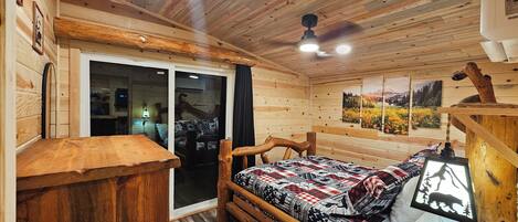 Ein Holzblockhaus-Interieur mit einem Bett, einem Holztisch, einer Schiebetür aus Glas und einem Deckenventilator.