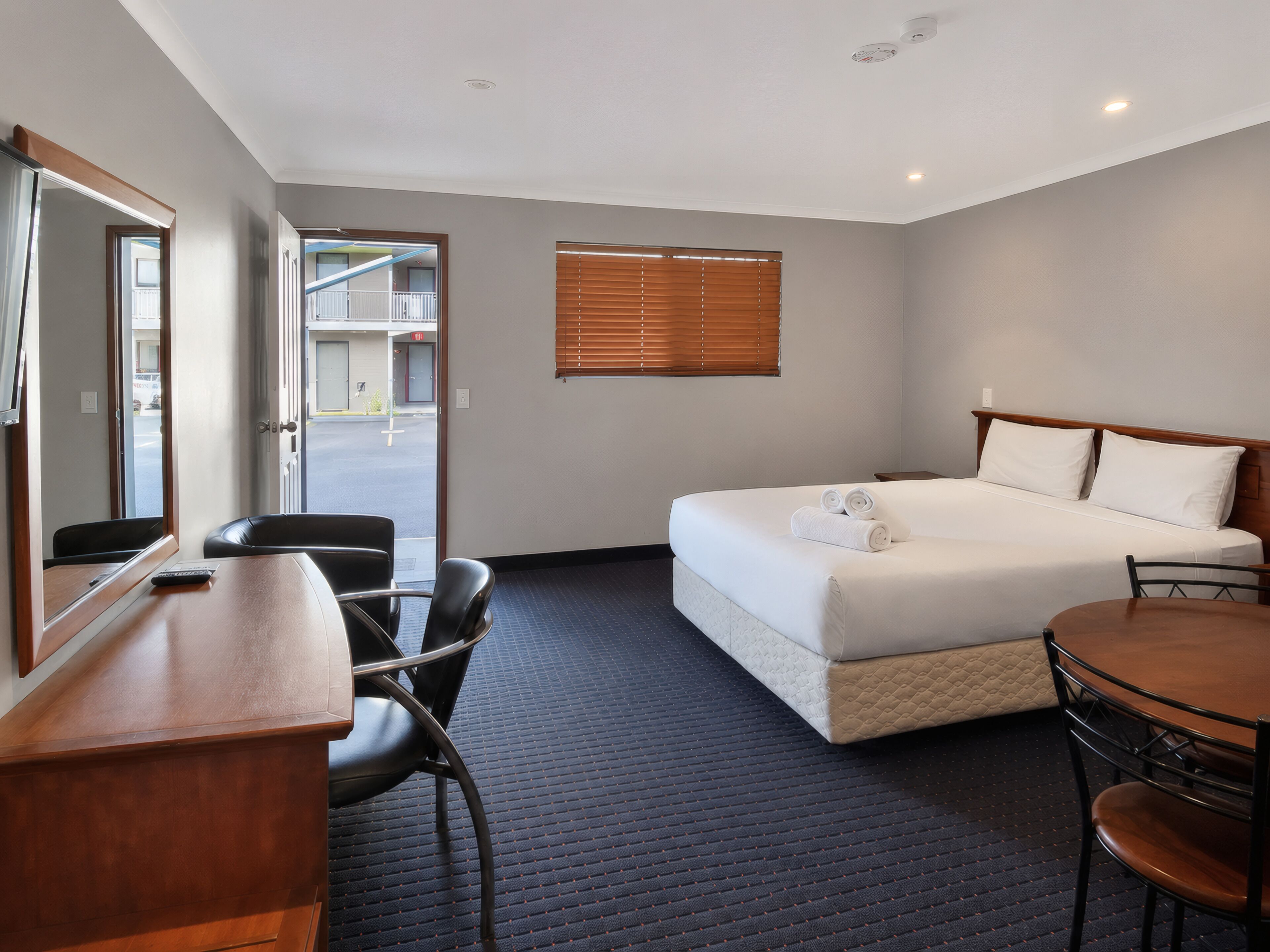 A hotel room with a bed, a desk, a chair, a window with blinds, and a view of the outside.