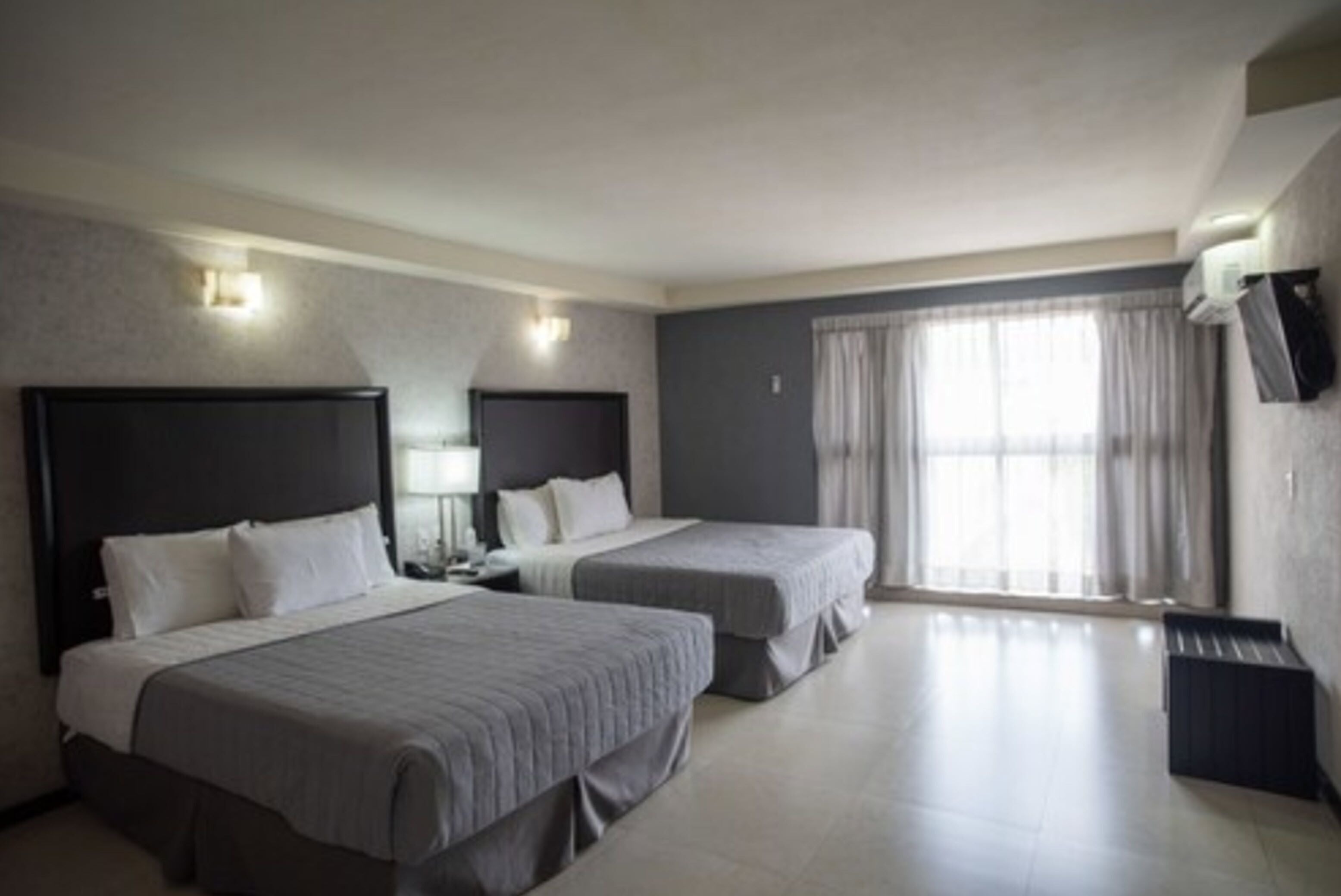 Una habitación de hotel con dos camas individuales, un televisor de pantalla plana, un ventanal grande con cortinas y un equipo de aire acondicionado integrado.