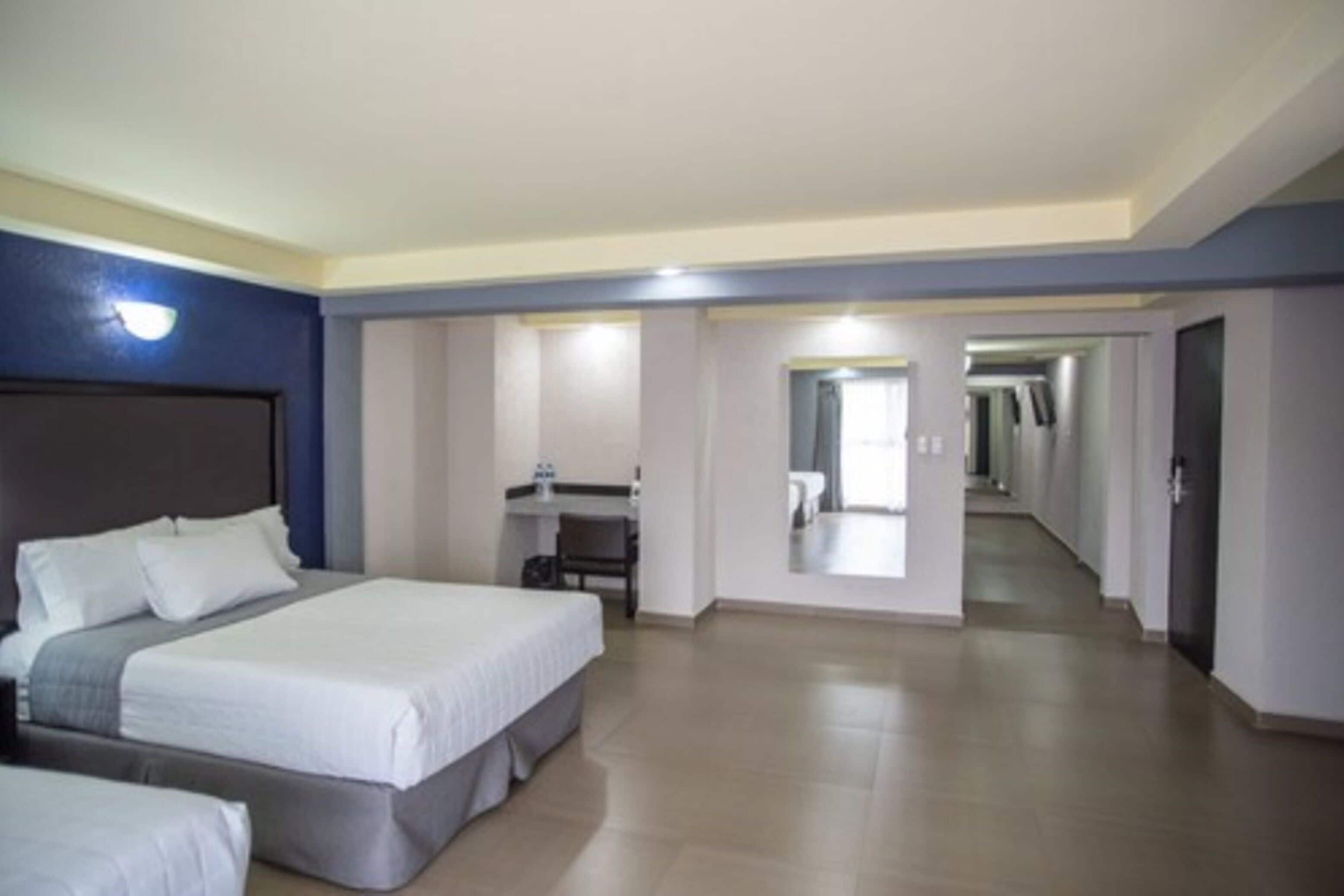 A hotel room with a large bed, a desk, and a mirror. There is a hallway leading to other rooms.