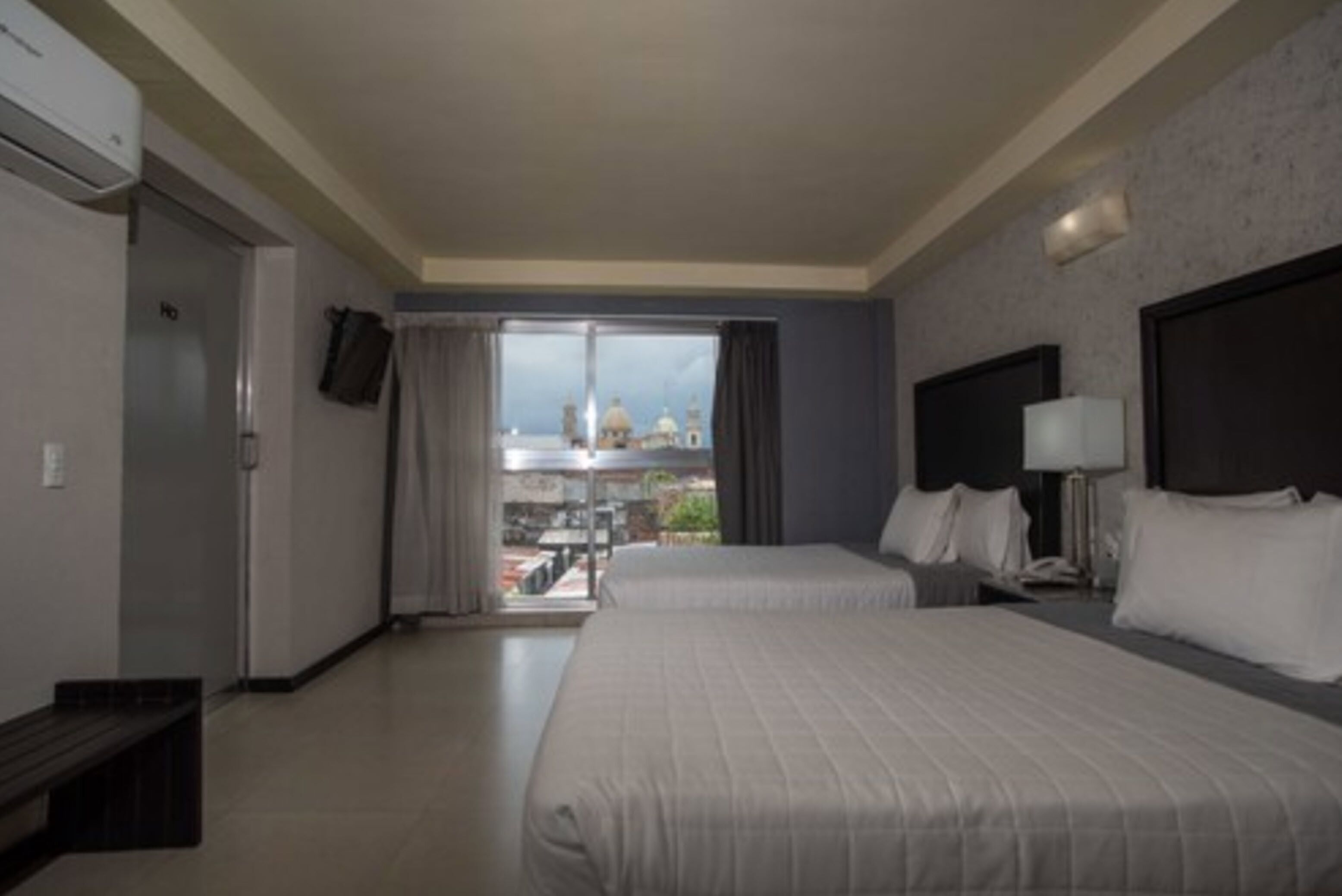 A hotel room with two beds, a large window with a view of buildings, a flat-screen TV, and a desk with a lamp.