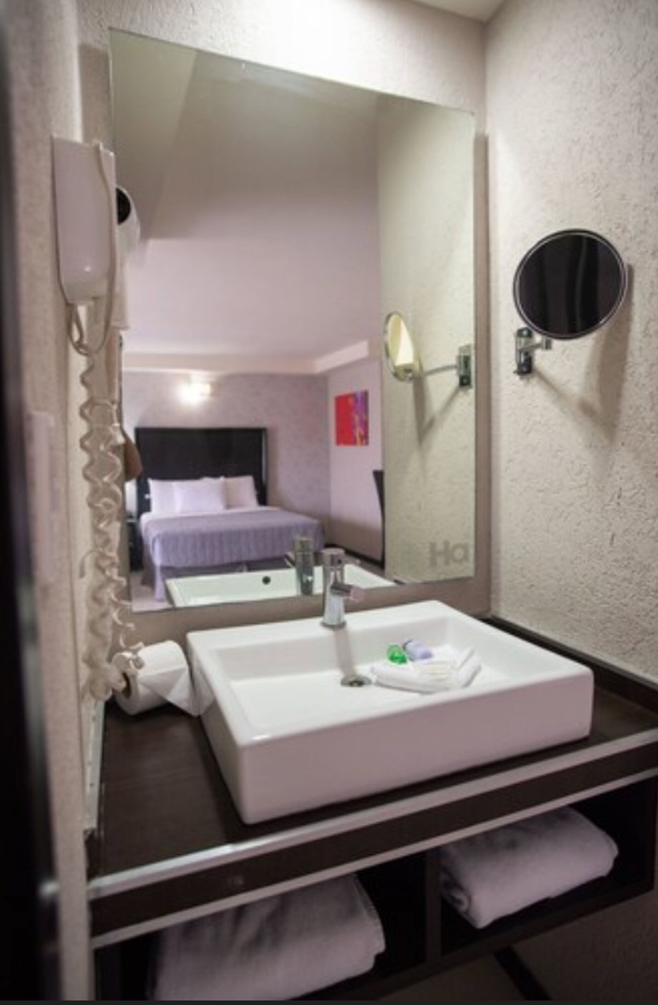 Un bagno d'albergo con un ampio specchio, un lavandino bianco, un asciugacapelli e un portascopevoli.