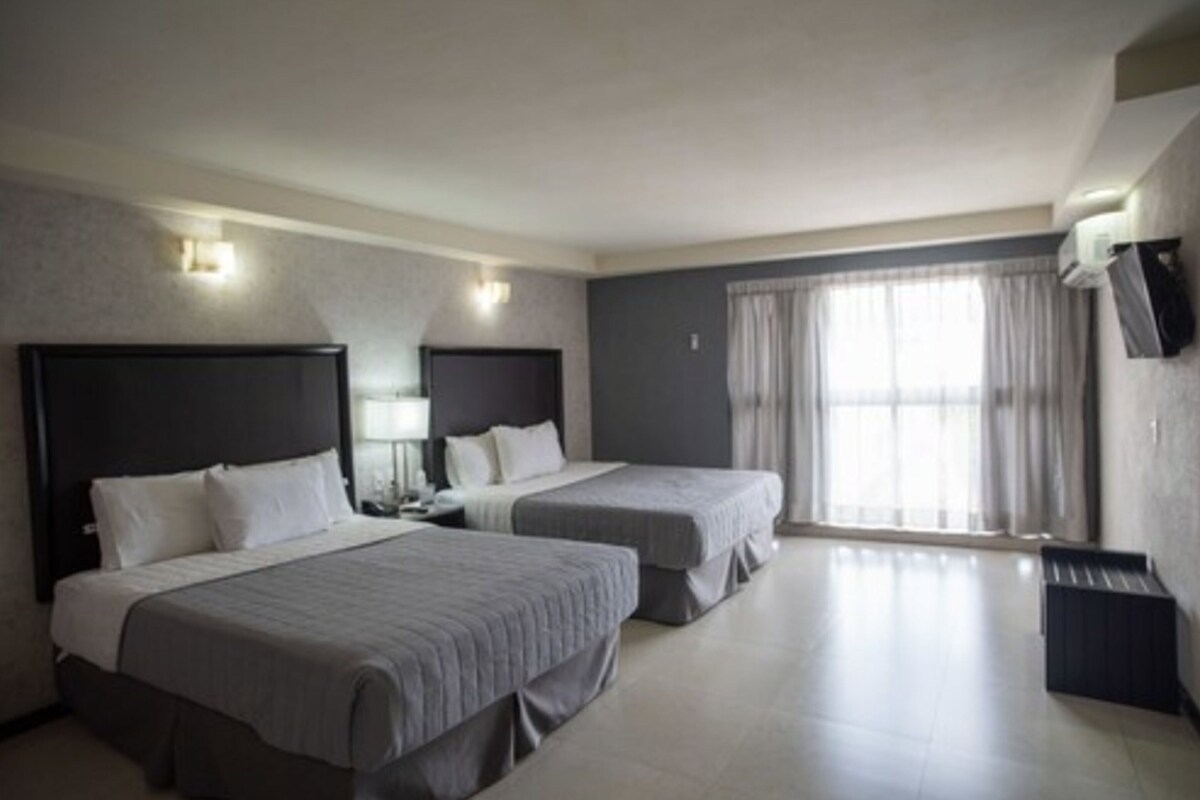 A hotel room with two beds, a flat-screen TV, a window with curtains, and a wall-mounted air conditioner.