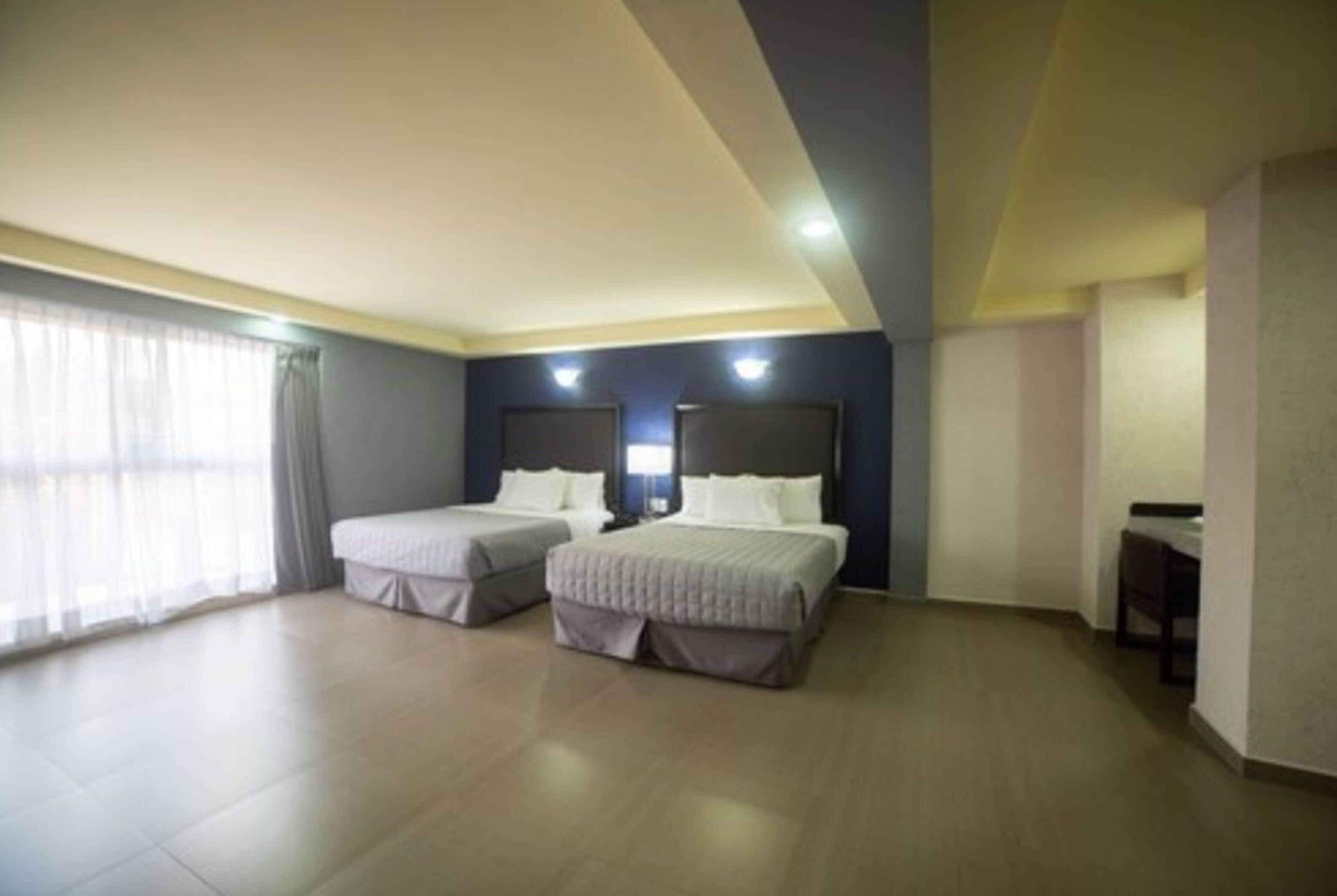 Quarto de hotel com duas camas, uma escrivaninha e uma janela grande com cortinas.