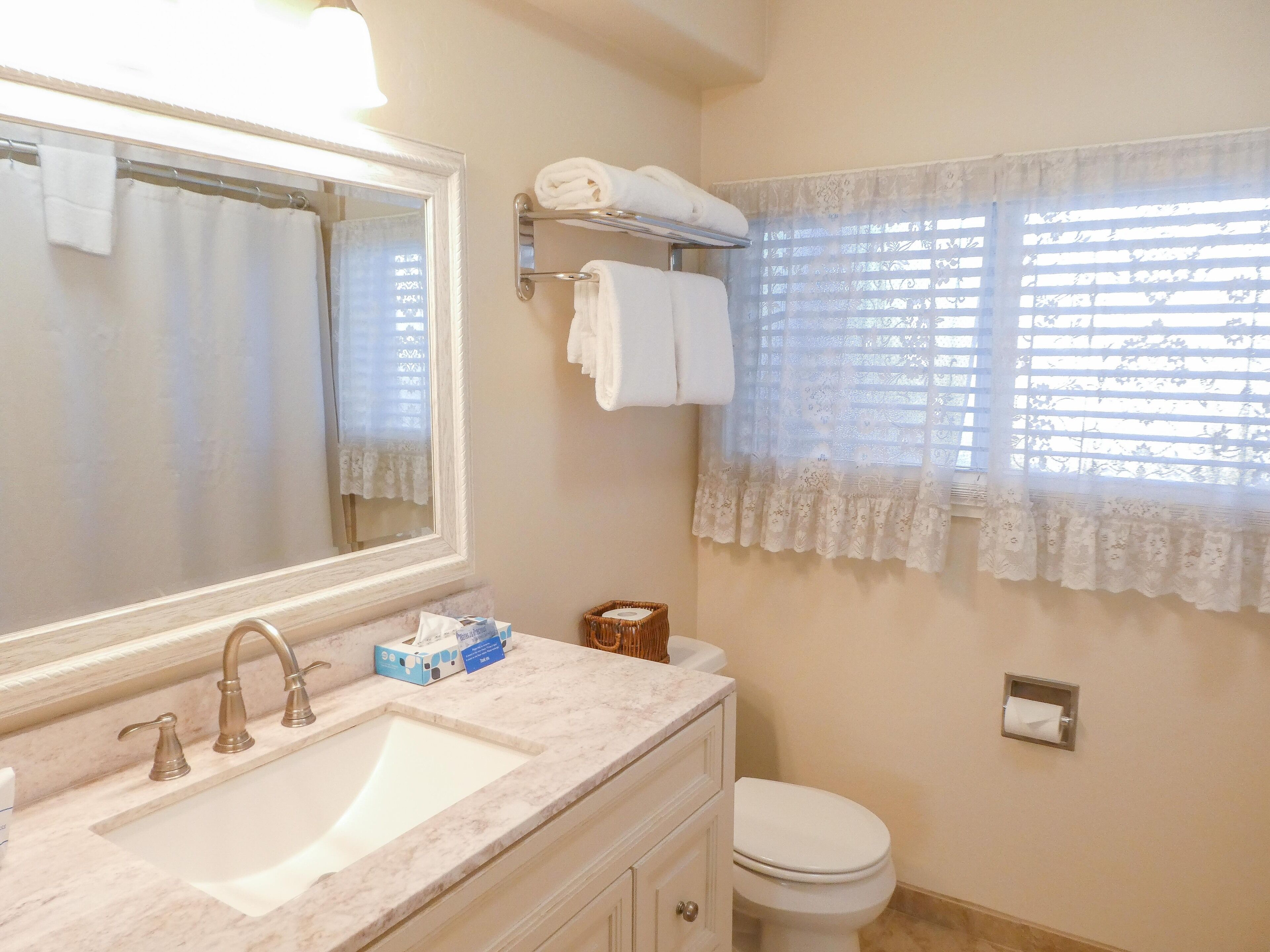 Baño con lavamanos, espejo, toallero, inodoro y ventana con cortinas de encaje.