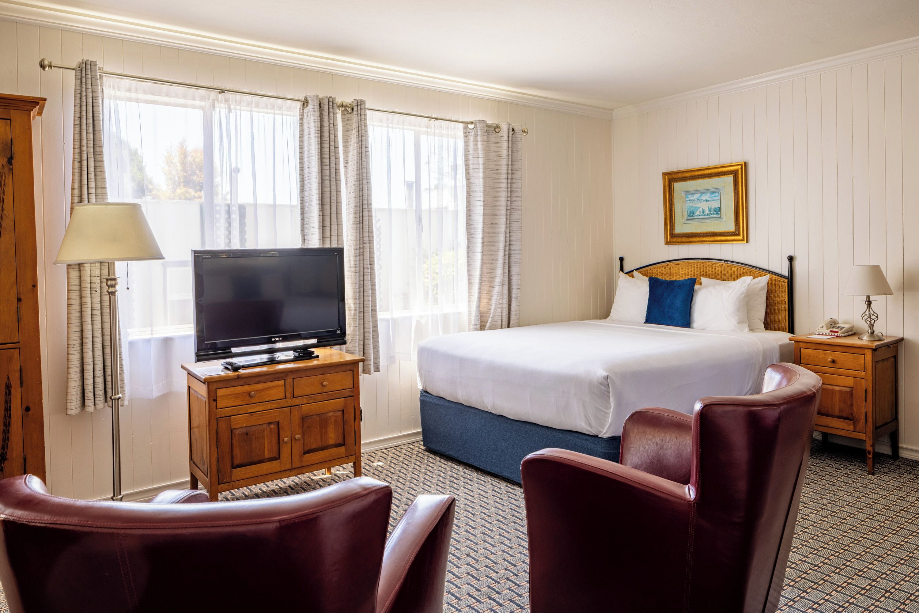 A hotel room with a bed, a television on a wooden cabinet, a desk with a lamp, and two leather chairs.