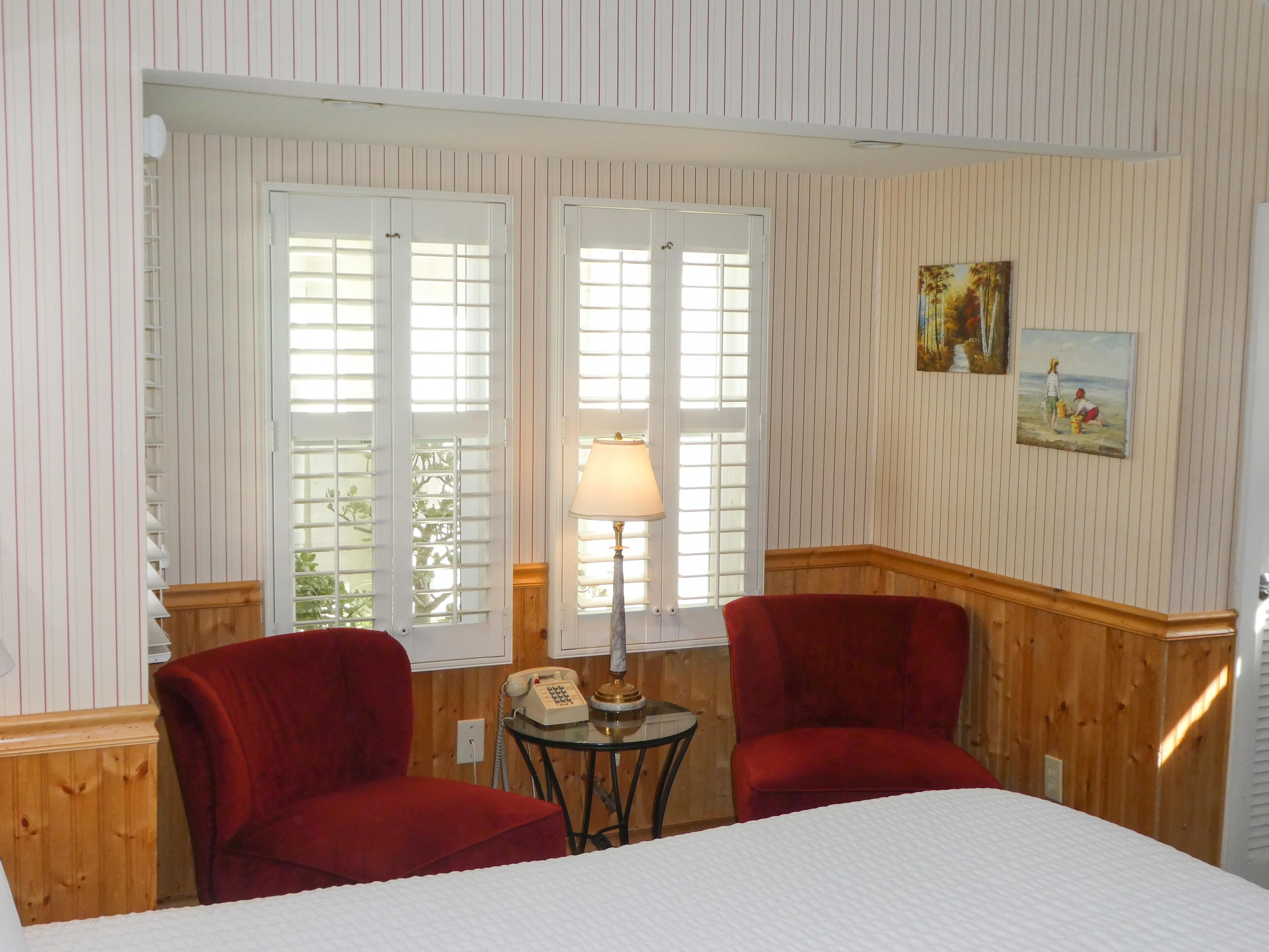 A hotel room with two red armchairs, a bed, a lamp, and two windows with shutters.