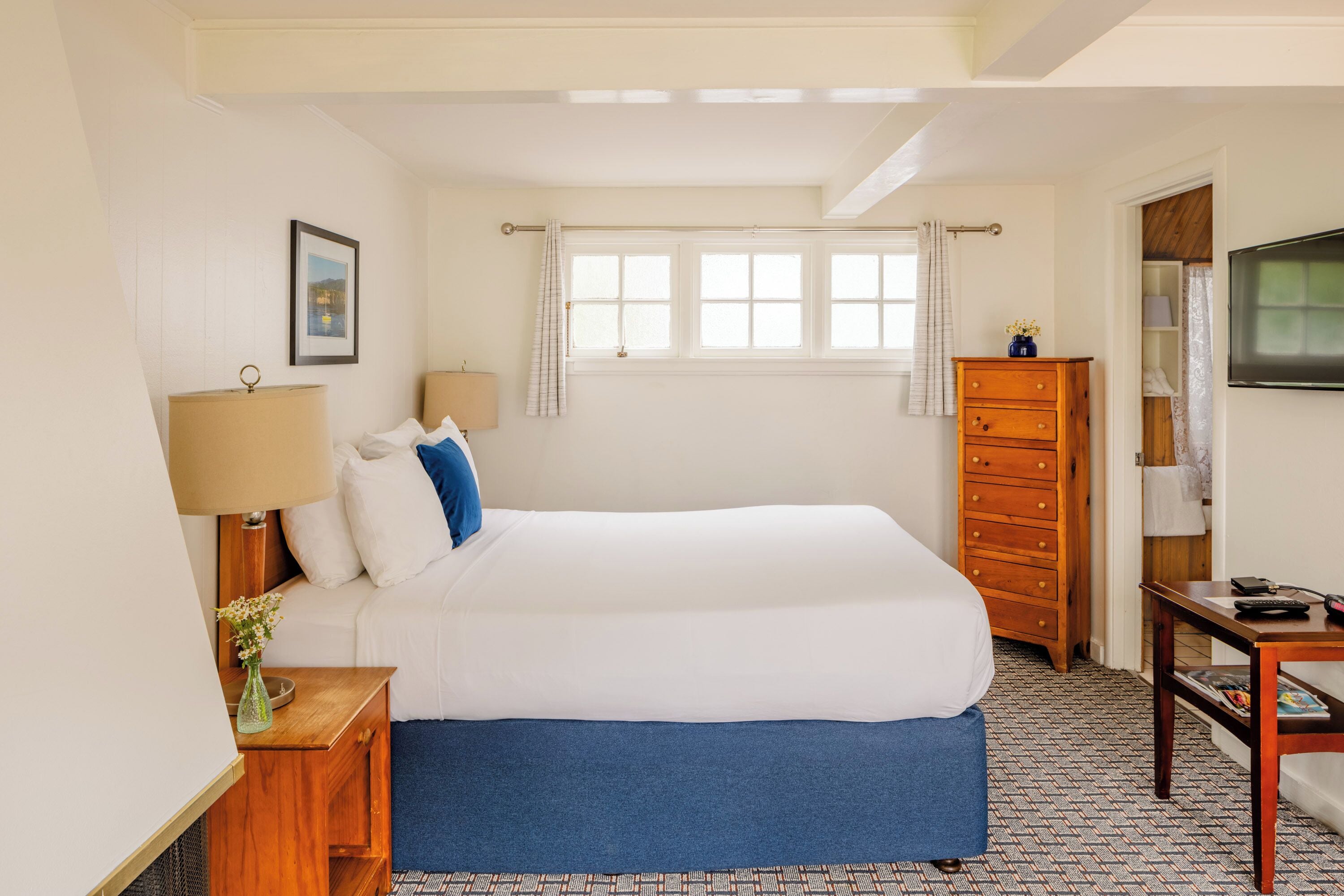 Una habitación de hotel con una cama, un aparador de madera, un escritorio con una silla, una lámpara, un cuadro en la pared y una ventana con cortinas.