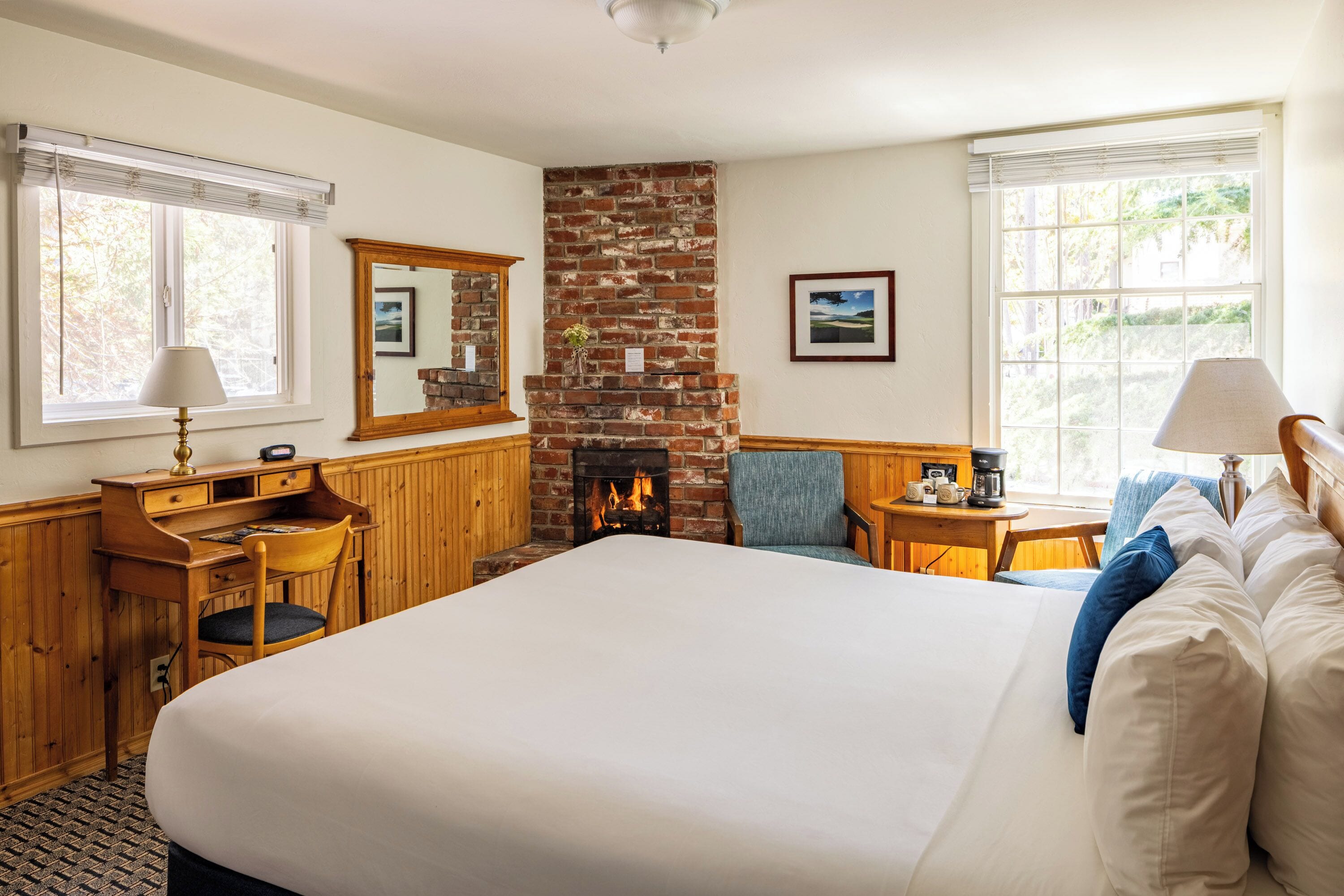A hotel room with a large bed, a wooden desk, a chair, a small table with a lamp, a fireplace, and a large window.