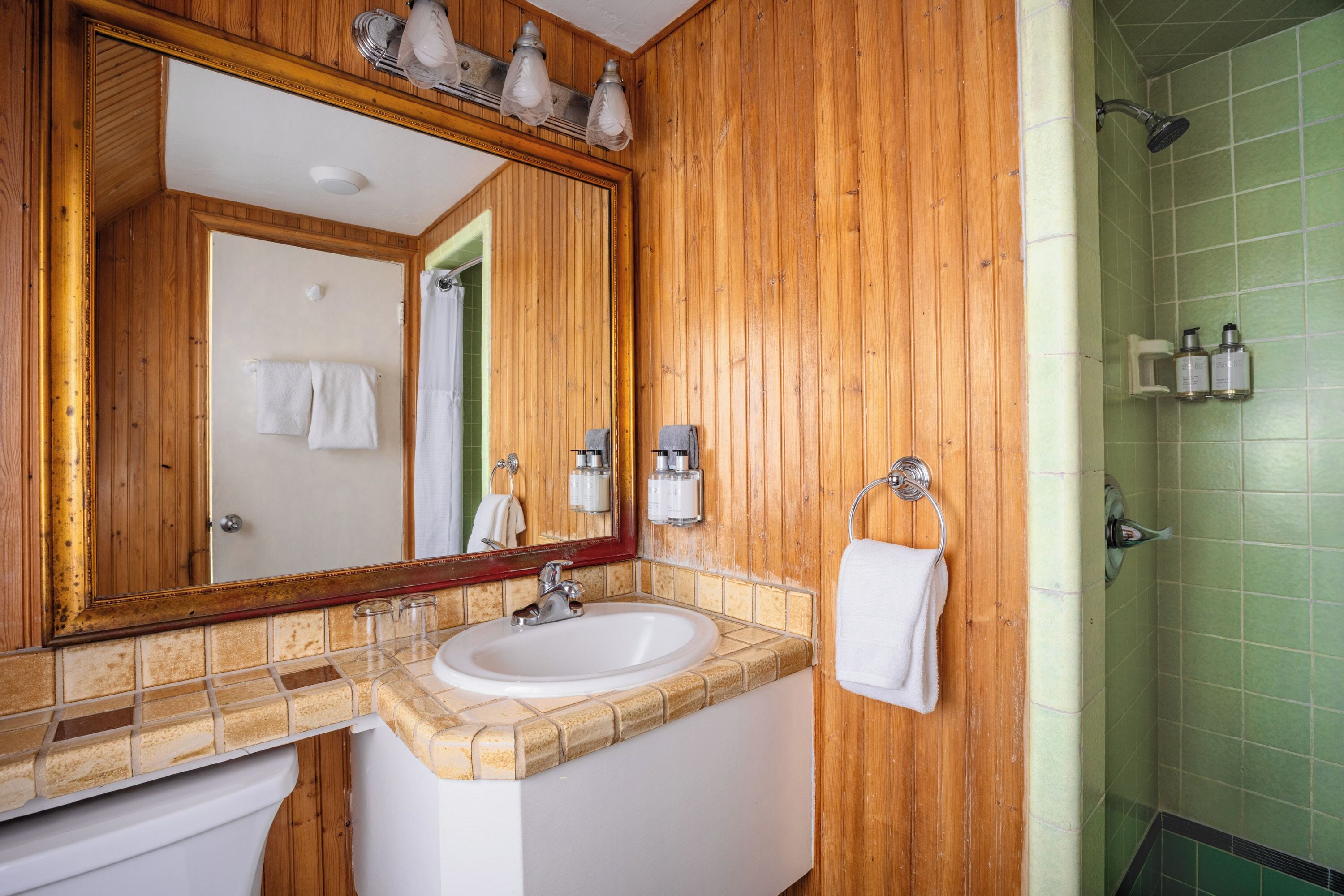 A bathroom with wooden paneling, a white sink, a large mirror, and a green-tiled shower area.