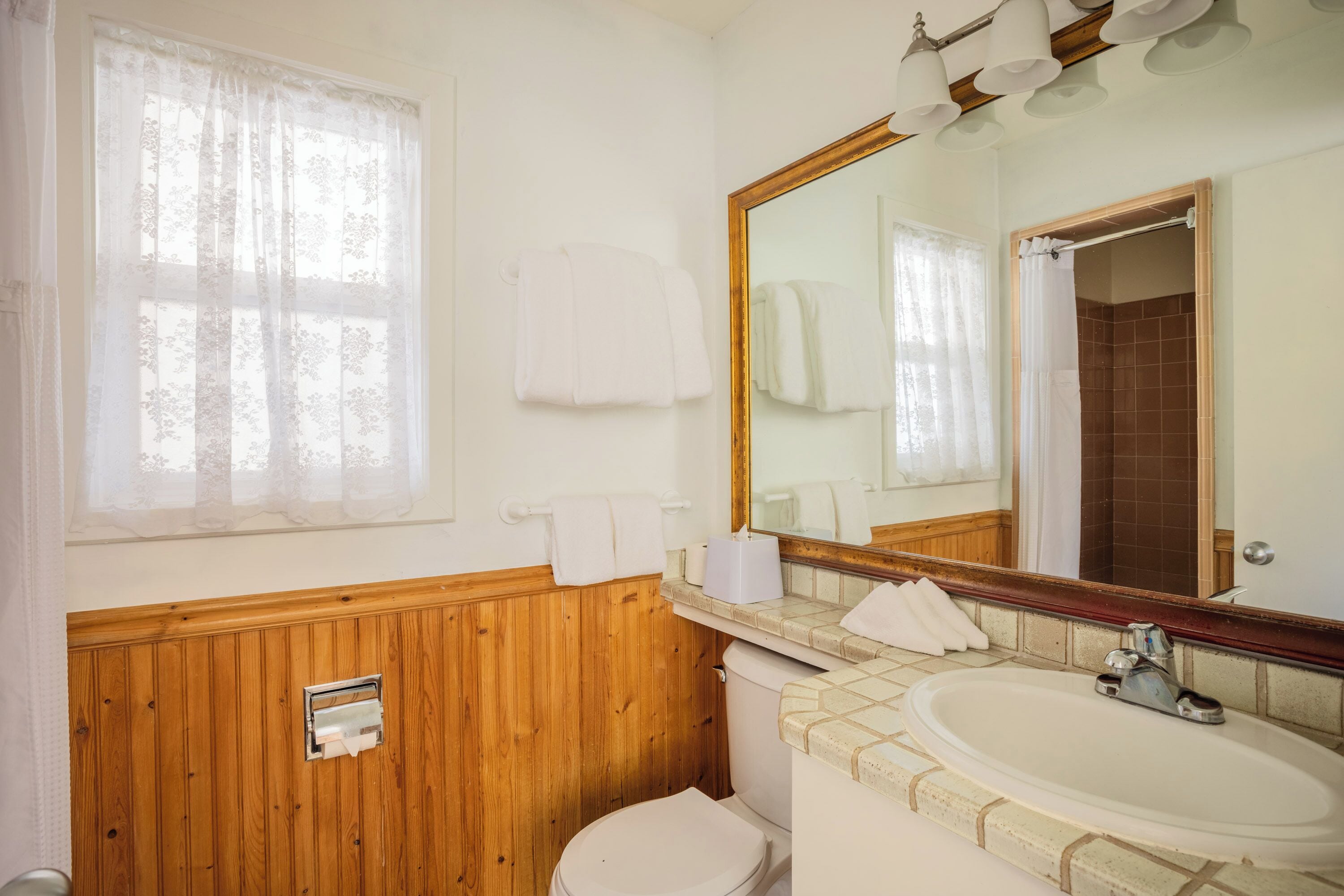 Een badkamer met een wand van houtpanelen, een wit toilet, een wastafel met spiegel en een raam met dunne gordijnen.