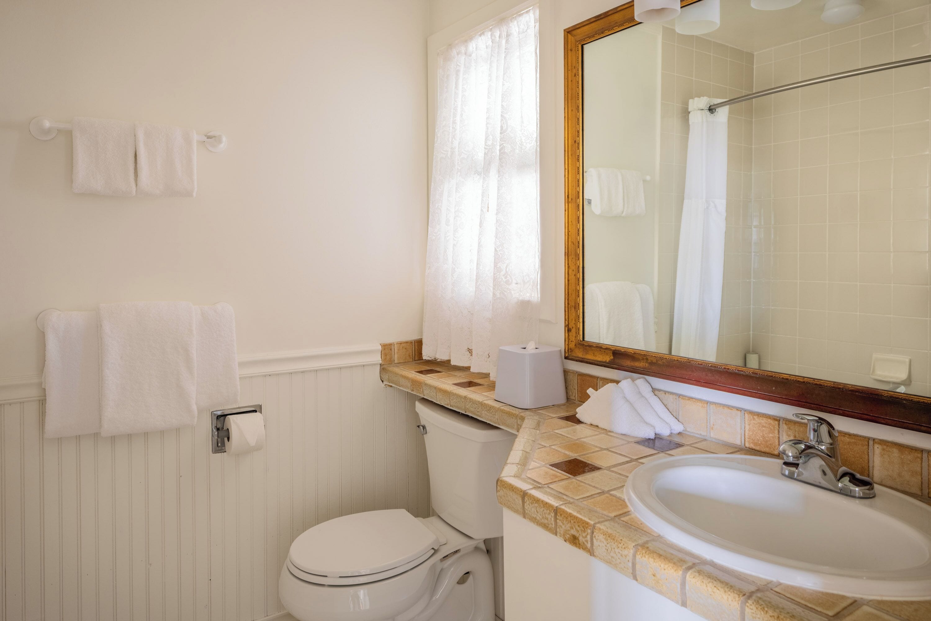 A bathroom with a toilet, sink, and mirror. There are towels hanging on the wall and a window with curtains.