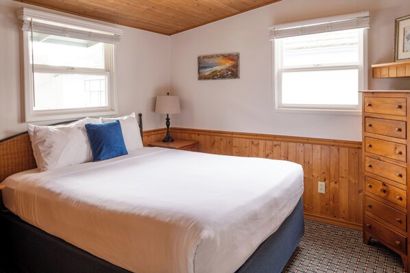 A bedroom with a large bed, wooden paneling, two windows with blinds, a nightstand with a lamp, and a dresser.