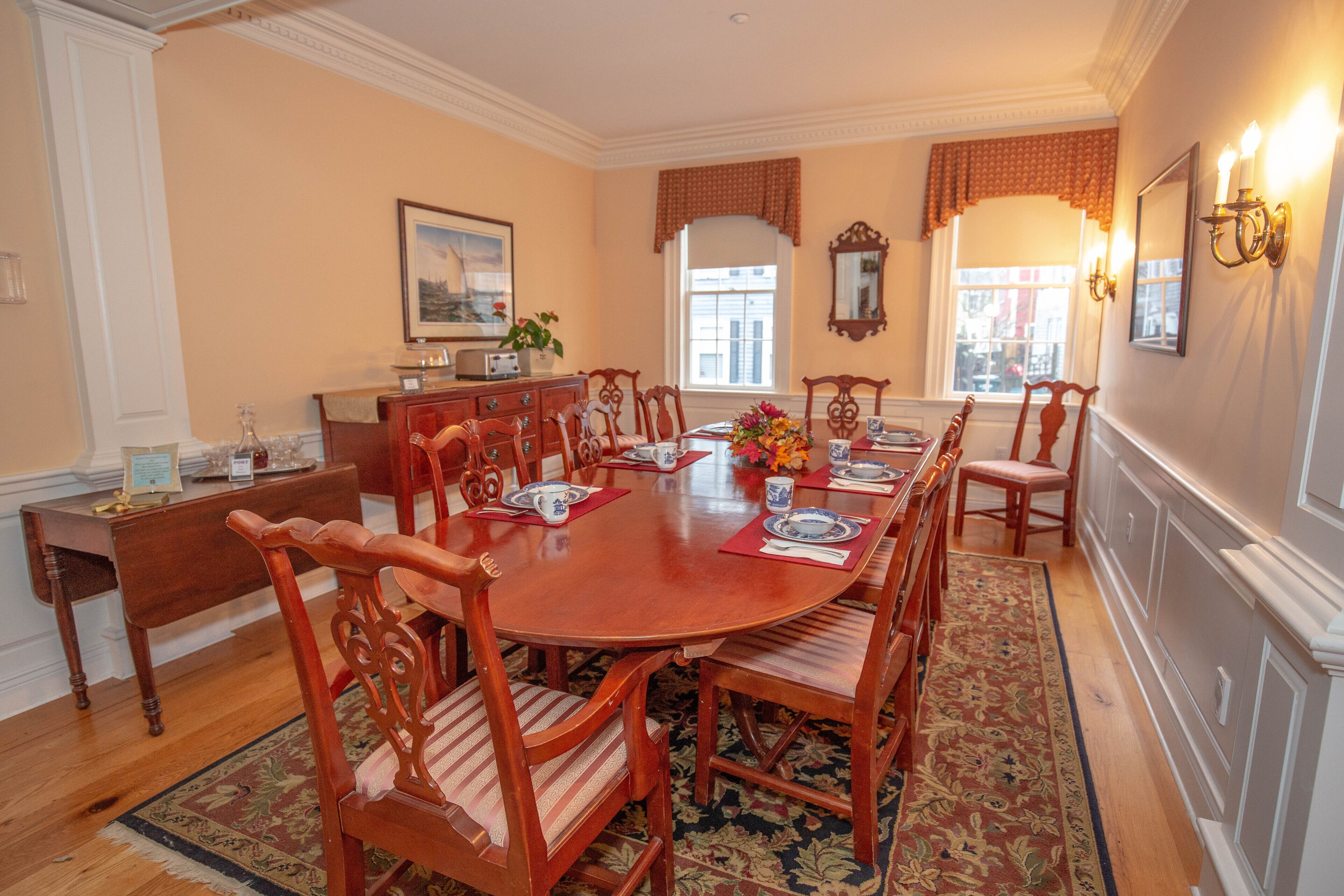 Une salle à manger avec une table ronde en bois, six chaises, un centre de table floral et un tapis à motifs.