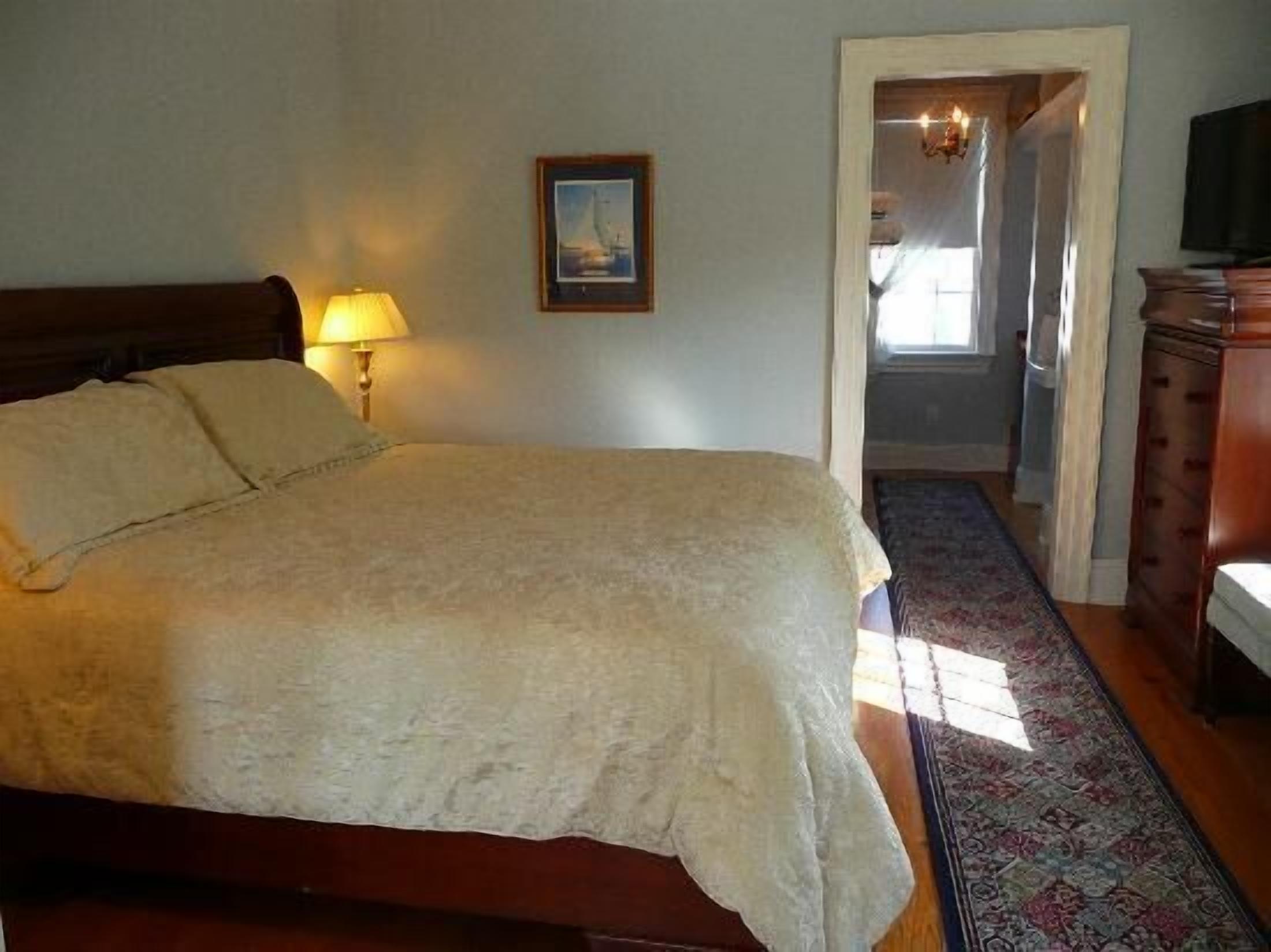 A bedroom with a large bed, a nightstand with a lamp, a framed picture on the wall, and a wooden dresser.