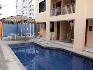 Una piscina con zona de estar cubierta por techo de paja, una escalera y un edificio de dos pisos con balcones.