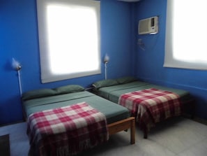 Una habitación con dos camas, cada una con una colcha verde y una manta a cuadros rojos y blancos. Se ve una ventana con persianas blancas, un equipo de aire acondicionado en la pared y una mesita con una lámpara.