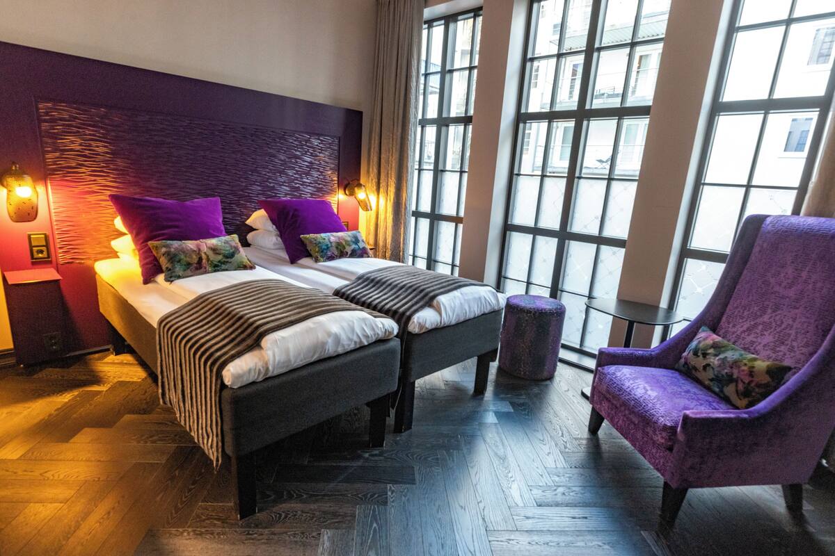 Habitación de hotel moderna con una cama grande, dos mesitas de noche, un sillón púrpura y vistas amplias a través de ventanas grandes.