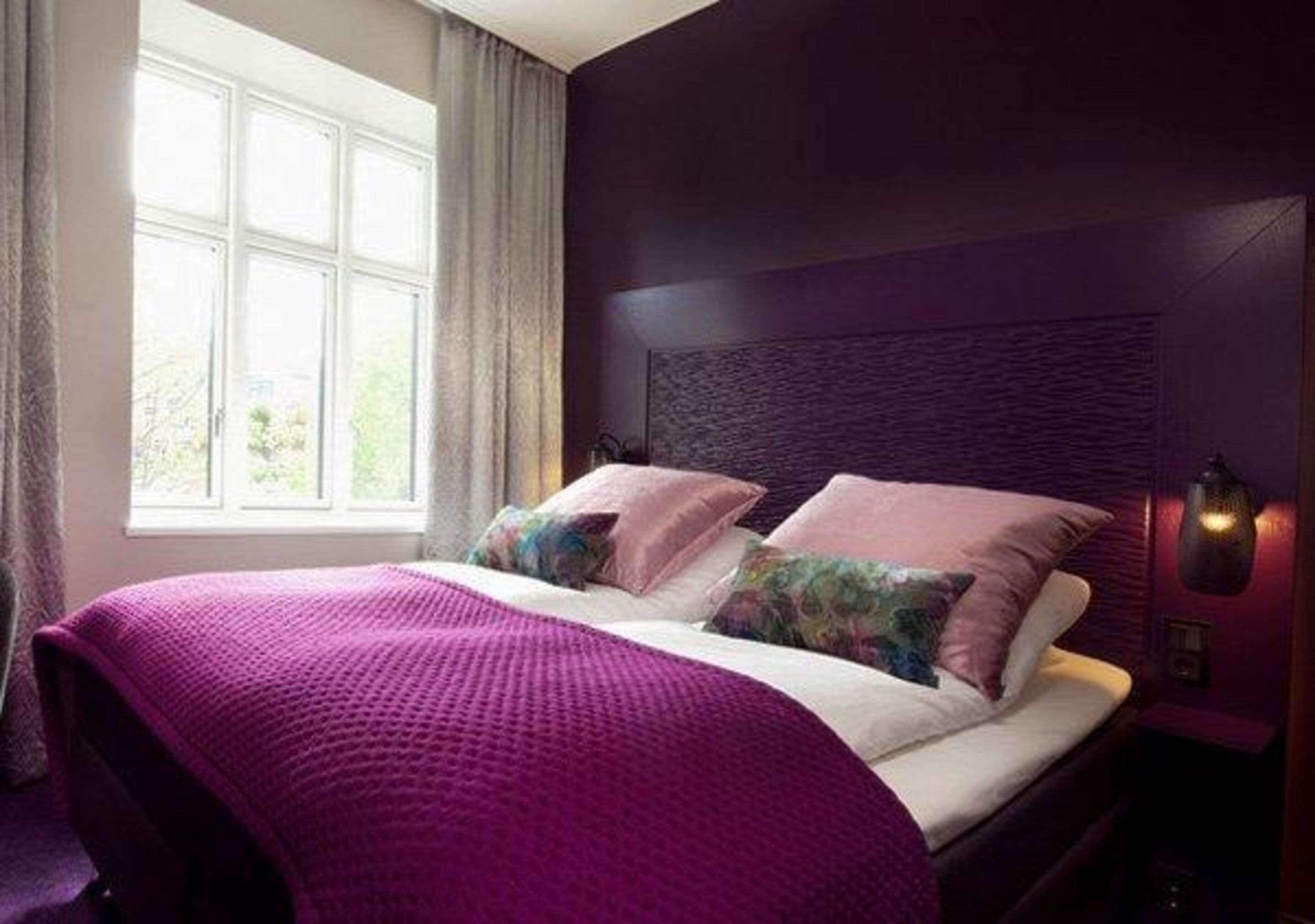 Un dormitorio con un ventanal grande, una cama con una colcha morada y una lámpara de pared.